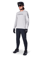 Alpinestars A-Dura Switch Long Sleeve Jersey, Light Gray, worn by male rider with black mountain bike pants and helmet, featuring black wordmark logo on chest and star graphics on sleeves, performance off-road cycling gear