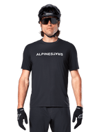 Mountain bike rider wearing the Alpinestars A-Dura Switch Short Sleeve Jersey in Black, featuring a white wordmark logo on the chest, styled with a black helmet, large sunglasses, and black gloves, professional cycling apparel for trail riding.