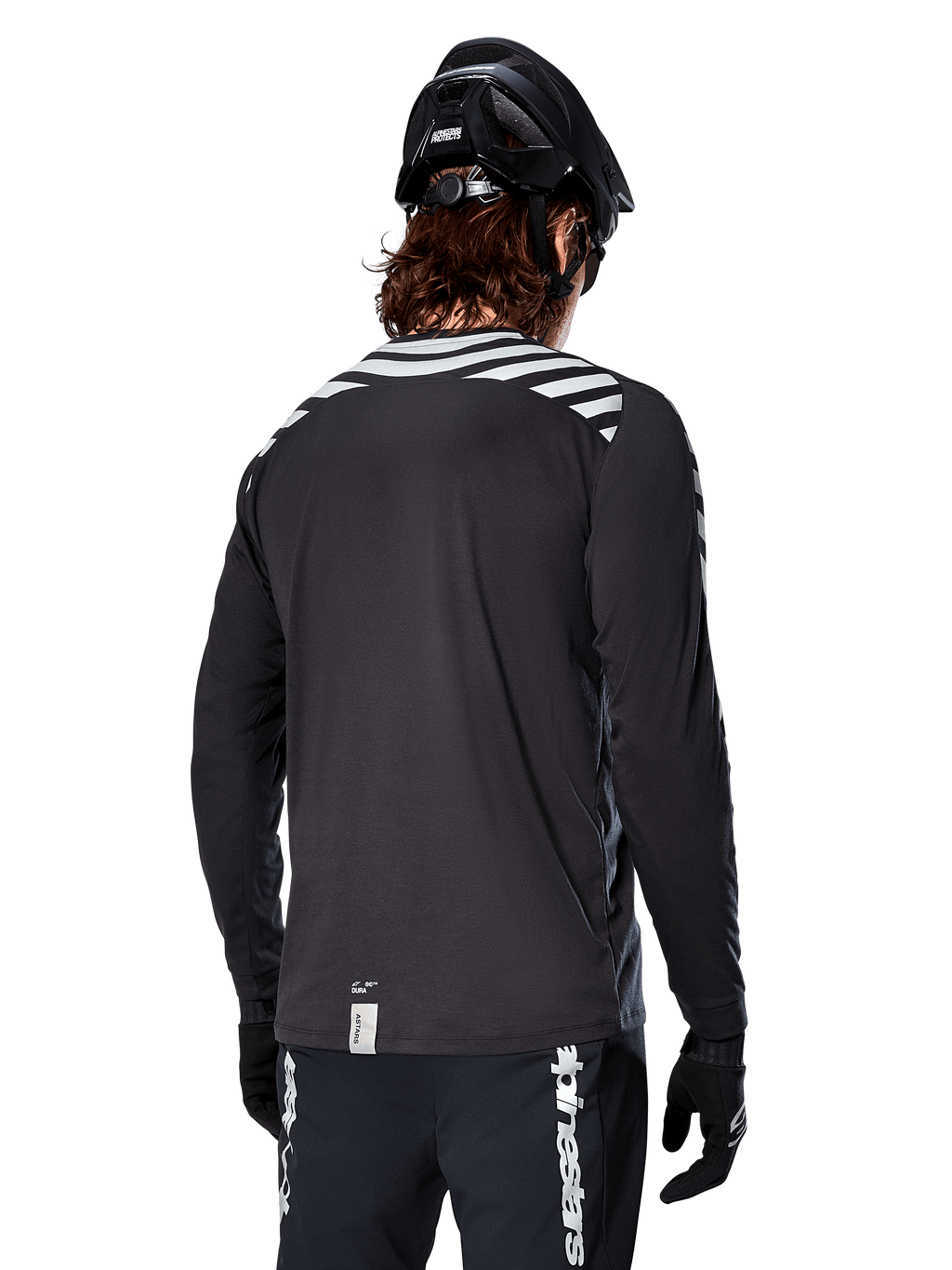 Alpinestars A-Dura Dri Oscar Langarmtrikot, Black Reflective, Rückansicht mit reflektierenden Diagonalstreifen auf den Schultern und einer verlängerten Rückenpartie, getragen mit schwarzem MTB-Helm und Handschuhen, technische Ausrüstung für Mountainbiking
