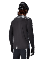 Alpinestars A-Dura Dri Oscar Langarmtrikot, Black Reflective, Rückansicht mit reflektierenden Diagonalstreifen auf den Schultern und einer verlängerten Rückenpartie, getragen mit schwarzem MTB-Helm und Handschuhen, technische Ausrüstung für Mountainbiking