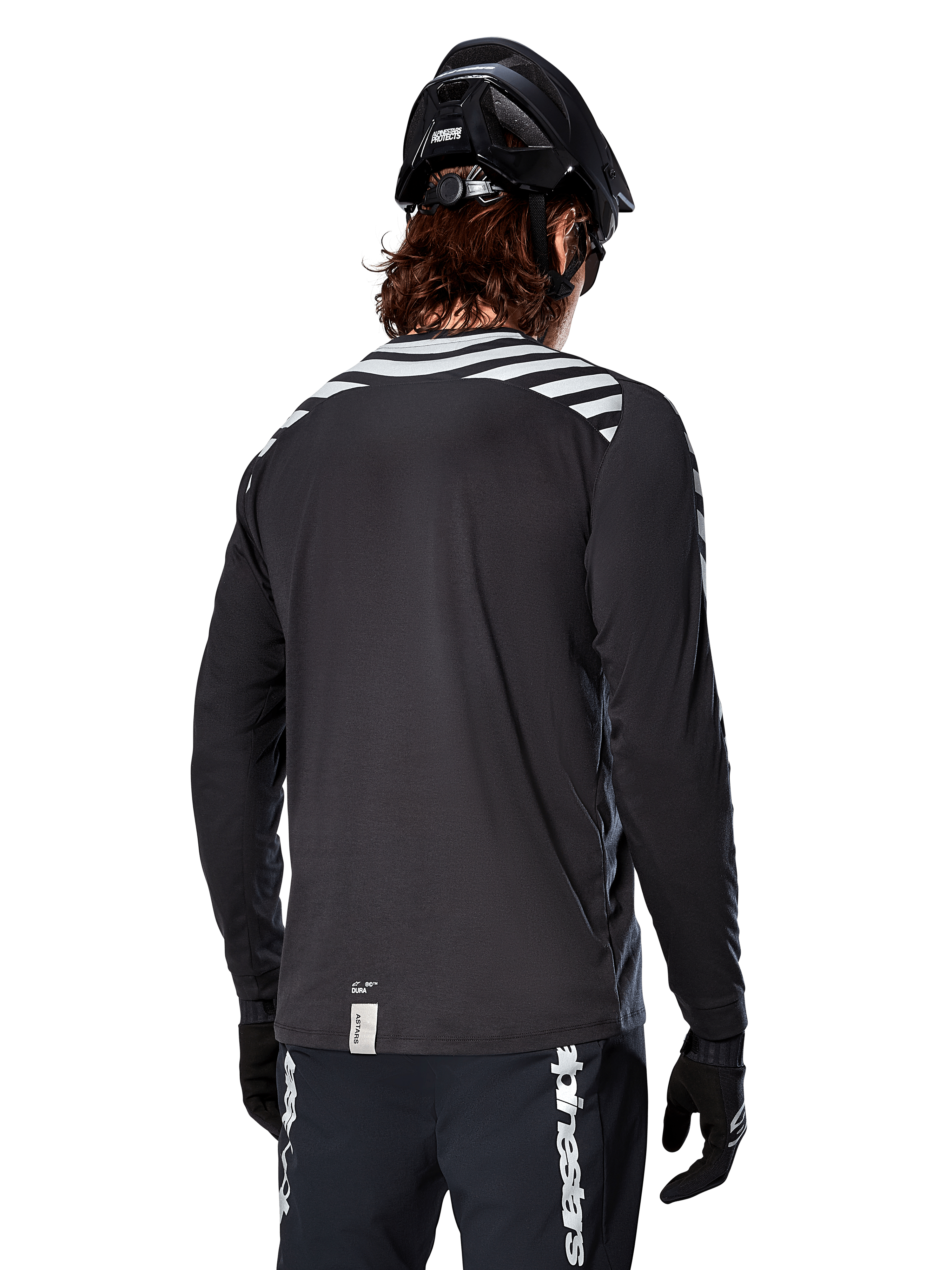 Alpinestars A-Dura Dri Oscar Langarmtrikot, Black Reflective, Rückansicht mit reflektierenden Diagonalstreifen auf den Schultern und einer verlängerten Rückenpartie, getragen mit schwarzem MTB-Helm und Handschuhen, technische Ausrüstung für Mountainbiking