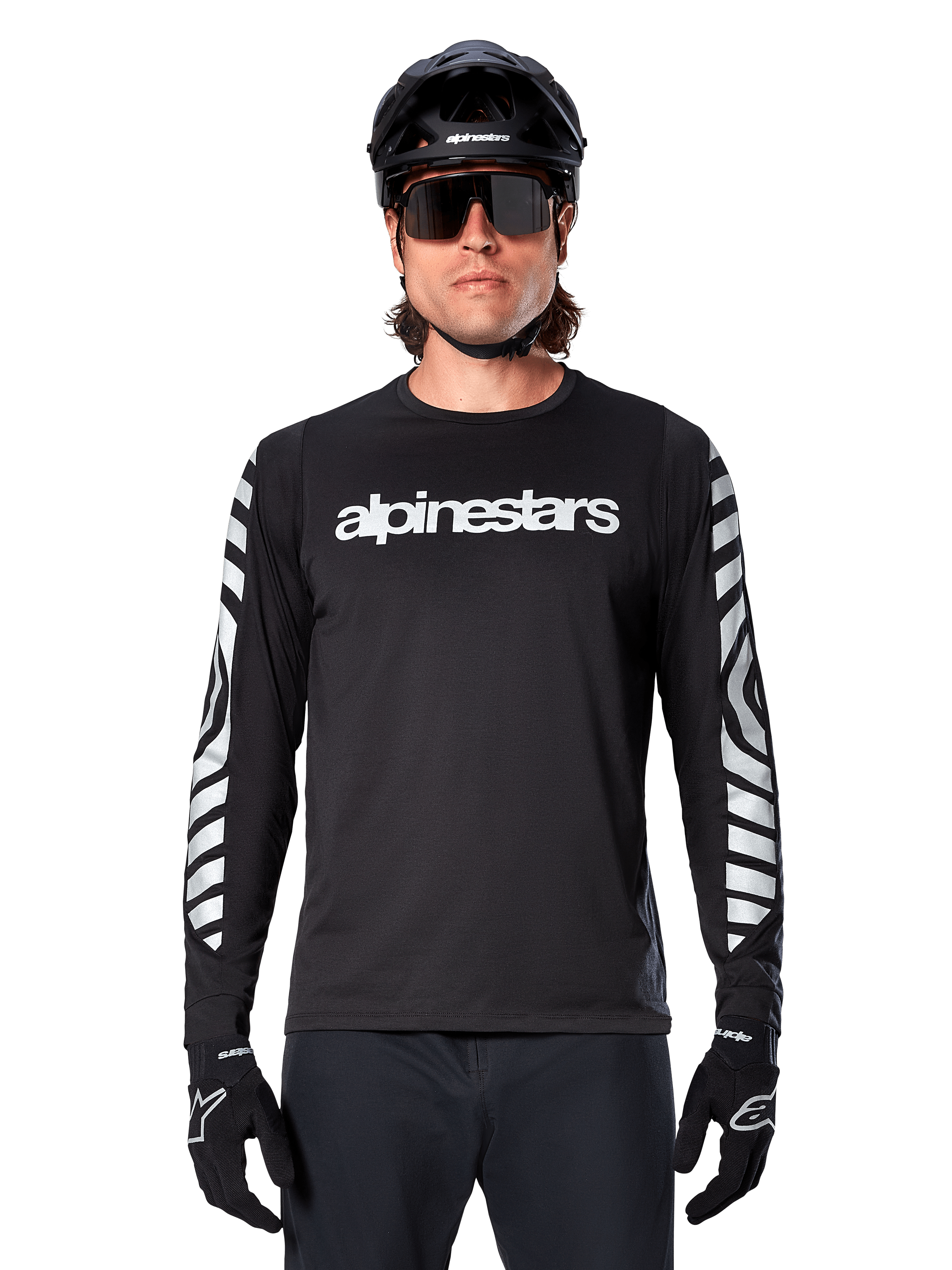 Männlicher Fahrer im Alpinestars A-Dura Dri Oscar Langarmtrikot, Schwarz Reflektierend, mit weißem Alpinestars-Schriftzug auf der Brust und silbernen reflektierenden Grafiken auf den Ärmeln, kombiniert mit schwarzem MTB-Helm, Sonnenbrille und schwarzen Handschuhen, Performance-Radsportbekleidung