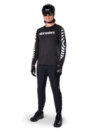 Alpinestars A-Dura Dri Oscar Langarmtrikot, Black Reflective, mit auffälligem weißem Alpinestars-Schriftzug auf der Brust und reflektierenden Grafikmustern auf den Ärmeln, kombiniert mit schwarzer Mountainbike-Hose, Helm und Handschuhen, technische MTB-Bekleidung