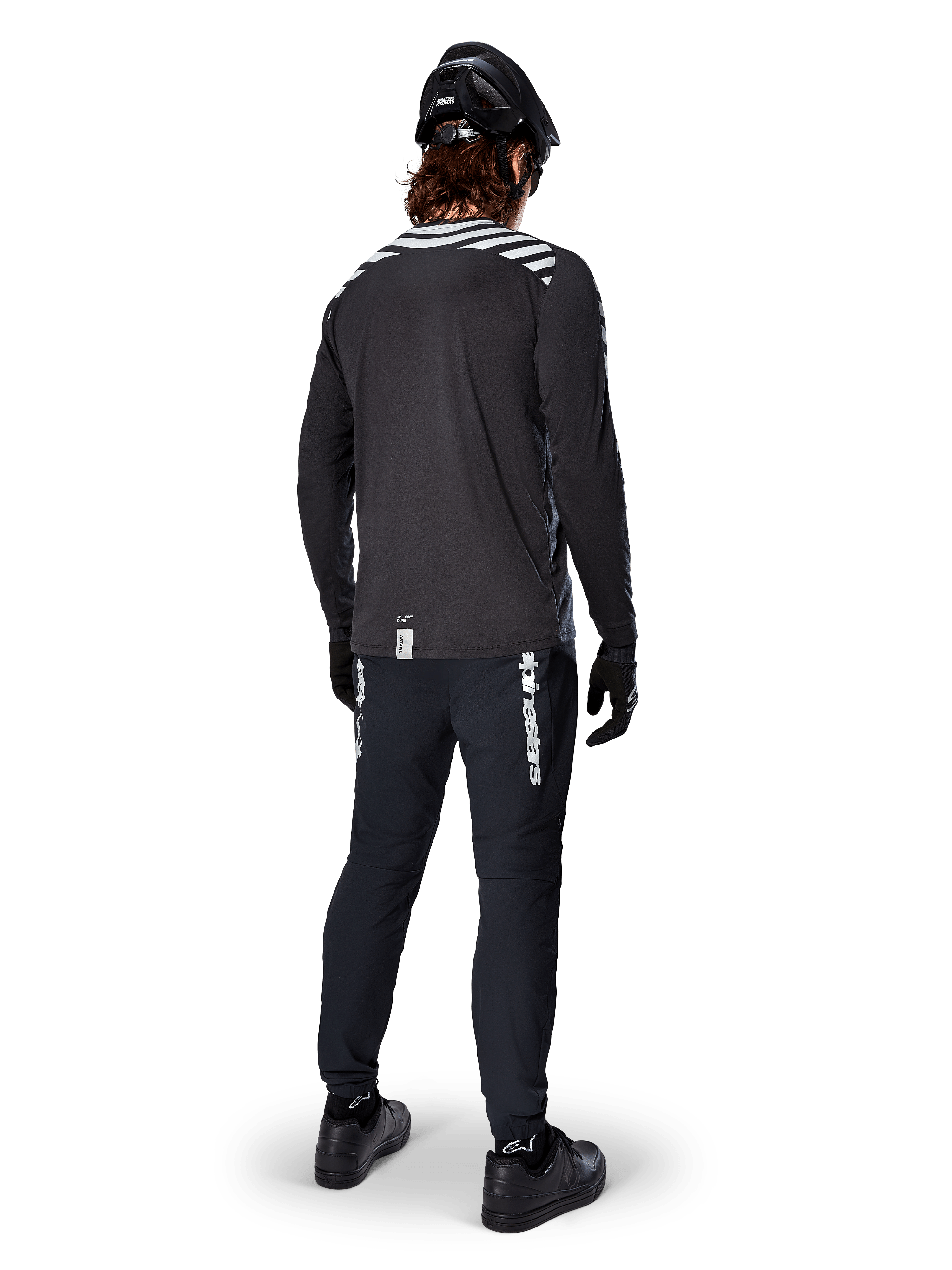Mountainbike-Fahrer im Alpinestars A-Dura Dri Oscar Langarmtrikot in Black Reflective, Rückansicht, kombiniert mit schwarzer technischer Hose, Helm und Handschuhen, mit reflektierenden Streifengrafiken auf den Schultern und einem verlängerten Rücken für den Offroad-Radsport.