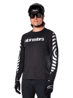 Alpinestars A-Dura Dri Oscar Langarmtrikot, MTB-Fahrradtrikot, Schwarz reflektierend, mit weißem Brust-Schriftzug und markanten reflektierenden kreisförmigen Grafiken auf den Ärmeln, getragen mit schwarzem Helm und Sonnenbrille, technische feuchtigkeitsableitende Bekleidung für Mountainbiking im Gelände