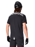 Alpinestars A-Dura Dri Oscar Trikot, Kurzarm-MTB-Trikot, Schwarz Reflektierend, Rückansicht am Model mit Fahrradhelm und Handschuhen, mit reflektierenden grauen Schulterstreifen und verlängertem Rückenteil, feuchtigkeitsableitendes Funktionsgewebe für Mountainbiking
