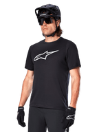 Alpinestars A-Dura Dri Oscar Trikot, Kurzarm-MTB-Radtrikot, Schwarz mit weißem Logo, feuchtigkeitsableitendes Funktionsmaterial, getragen mit schwarzem Helm, Sonnenbrille und Handschuhen