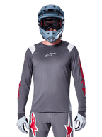 MTB rider wearing Alpinestars A-Supra Astar Long Sleeve Jersey, Dark Gray with red graphics on sleeves and white shoulder accents, matching gray pants with red star logos, blue full-face helmet and goggles, coordinated off-road cycling gear