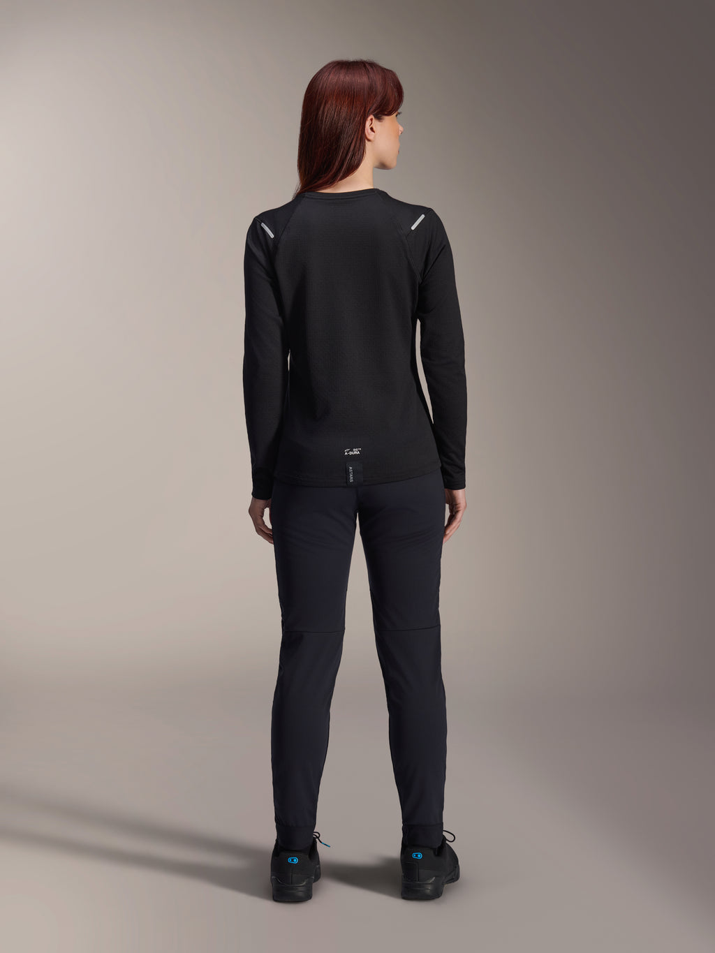 Alpinestars Damen Stella A-Dura Thermo-Trikot, Rückansicht, Schwarz, langärmliges Radtrikot mit isoliertem Fleece, getragen mit passender schwarzer Hose und Schuhen, mit kleinem A-Dura-Branding am unteren Rücken und reflektierenden Akzenten an den Schultern, speziell für Mountainbike-Fahrten von Frauen entwickelt.