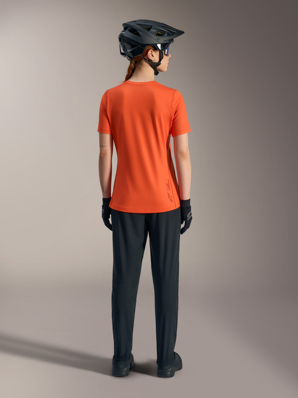 Alpinestars Women's Stella A-Dura Ride Short Sleeve Jersey in orange, back view with black MTB pants and helmet.