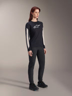 Frau in Alpinestars MTB-Ausrüstung, schwarzes Stella A-Dura Elite Trikot mit weißem Brustlogo und Ärmeleinsätzen, schwarze Stella A-Dura Hose, schwarze Fahrradschuhe mit blauen Akzenten, komplettes Performance-Outfit für das Mountainbiken