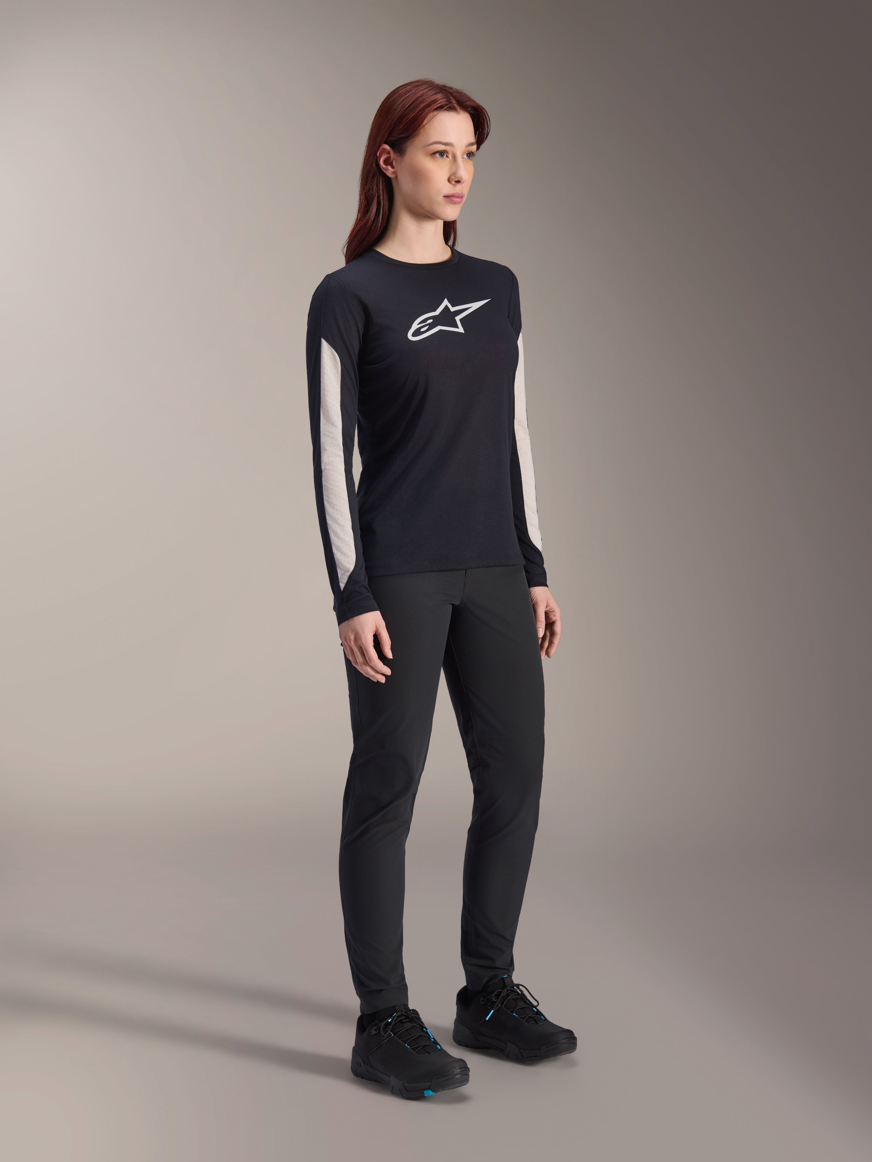Frau in Alpinestars MTB-Ausrüstung, schwarzes Stella A-Dura Elite Trikot mit weißem Brustlogo und Ärmeleinsätzen, schwarze Stella A-Dura Hose, schwarze Fahrradschuhe mit blauen Akzenten, komplettes Performance-Outfit für das Mountainbiken
