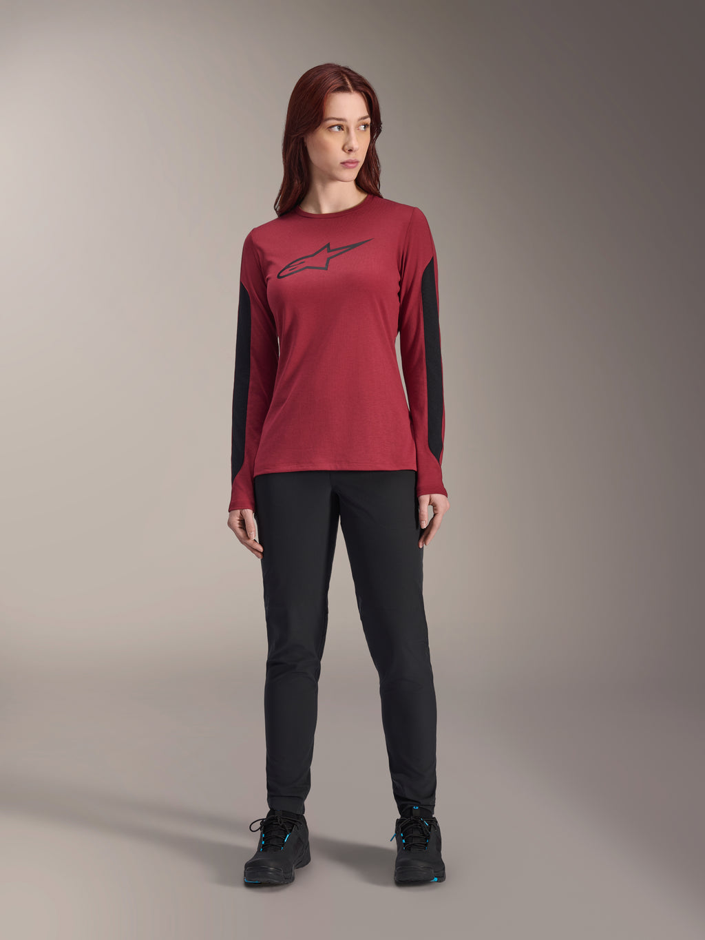 Alpinestars Stella A-Dura Dri Astar Langarmtrikot für Damen in Burgund, mit einem schwarzen Astars-Logo auf der Brust und schwarzen Mesh-Einsätzen an den Armen, kombiniert mit einer schwarzen technischen Hose und Sportschuhen, Performance-Mountainbike-Bekleidung für Damen.
