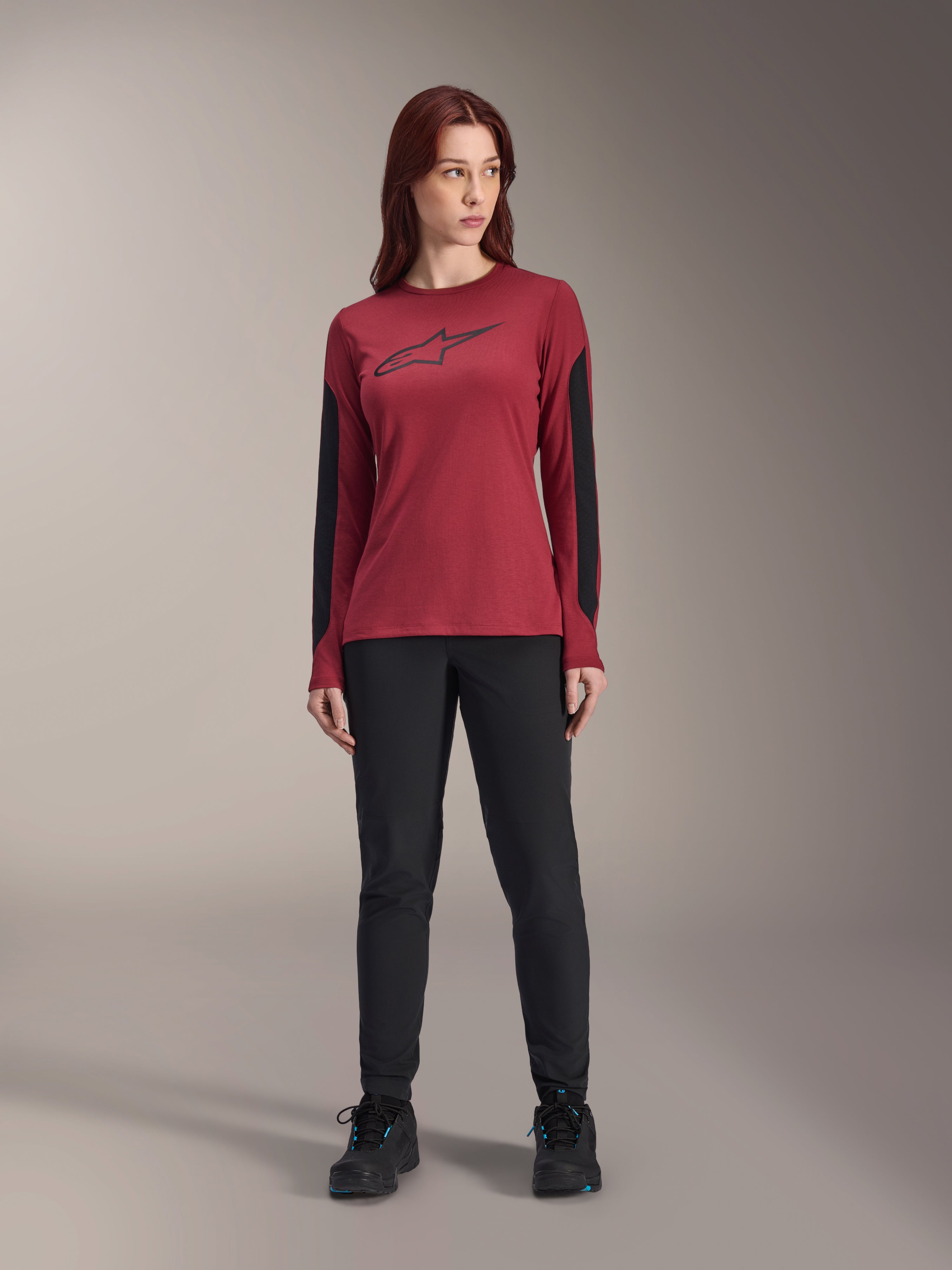 Alpinestars Stella A-Dura Dri Astar Langarmtrikot für Damen in Burgund, mit einem schwarzen Astars-Logo auf der Brust und schwarzen Mesh-Einsätzen an den Armen, kombiniert mit einer schwarzen technischen Hose und Sportschuhen, Performance-Mountainbike-Bekleidung für Damen.