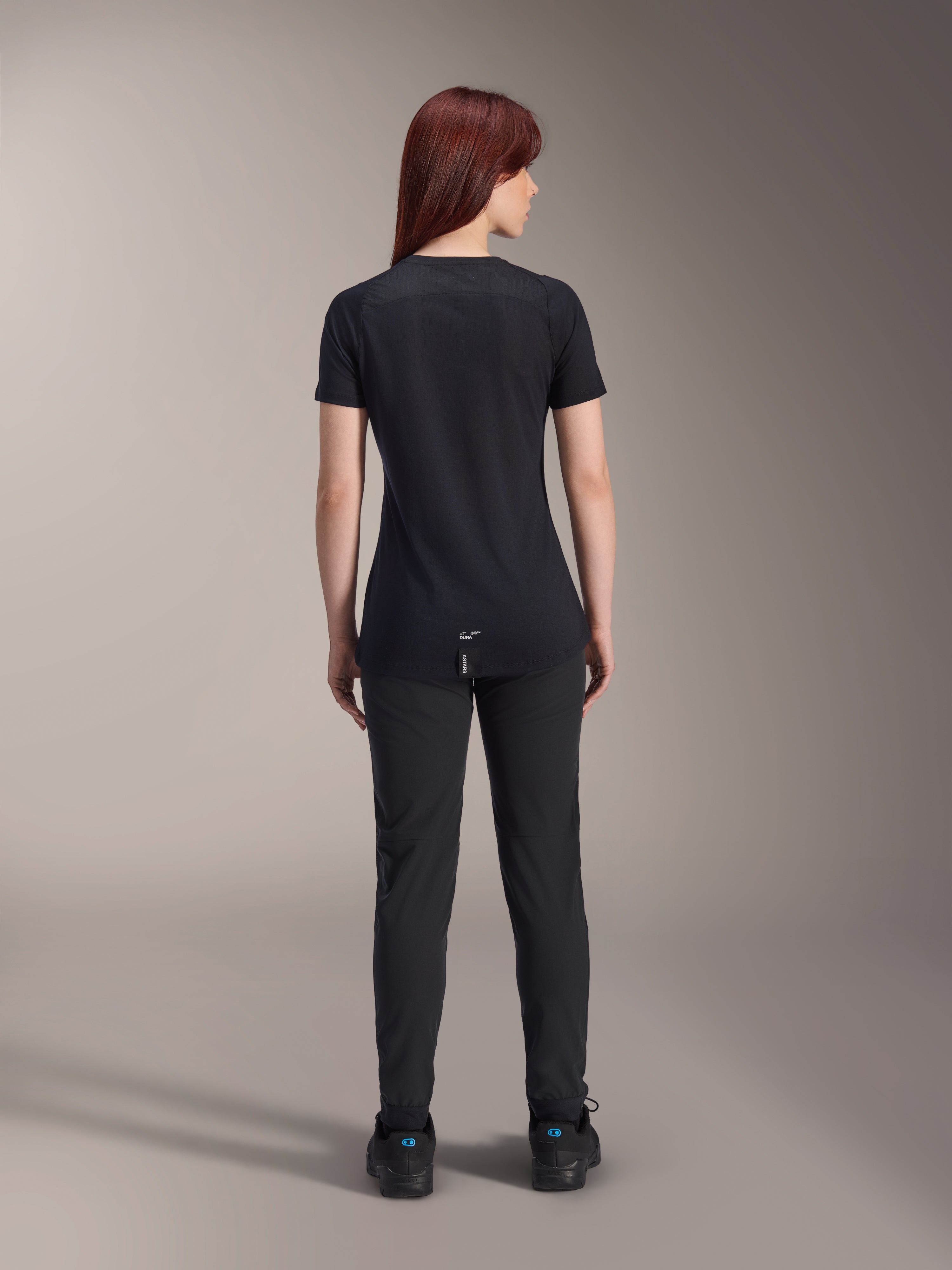 Alpinestars Damen Stella A-Dura Dri Switch Kurzarmtrikot, Schwarz, Rückansicht an einem Model, mit längerem abfallendem Saum und technischen Mesh-Einsätzen, kombiniert mit schwarzer Hose, Performance-MTB-Fahrradausrüstung
