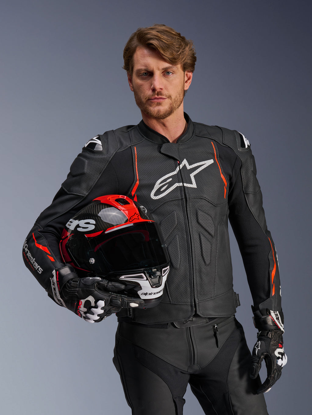 Alpinestars Dusk Airflow Leather Jacket, Black with Red Fluo accents, worn by a male model holding a red and black motorcycle helmet, featuring perforated leather for maximum ventilation and white Alpinestars branding, sport motorcycle riding gear