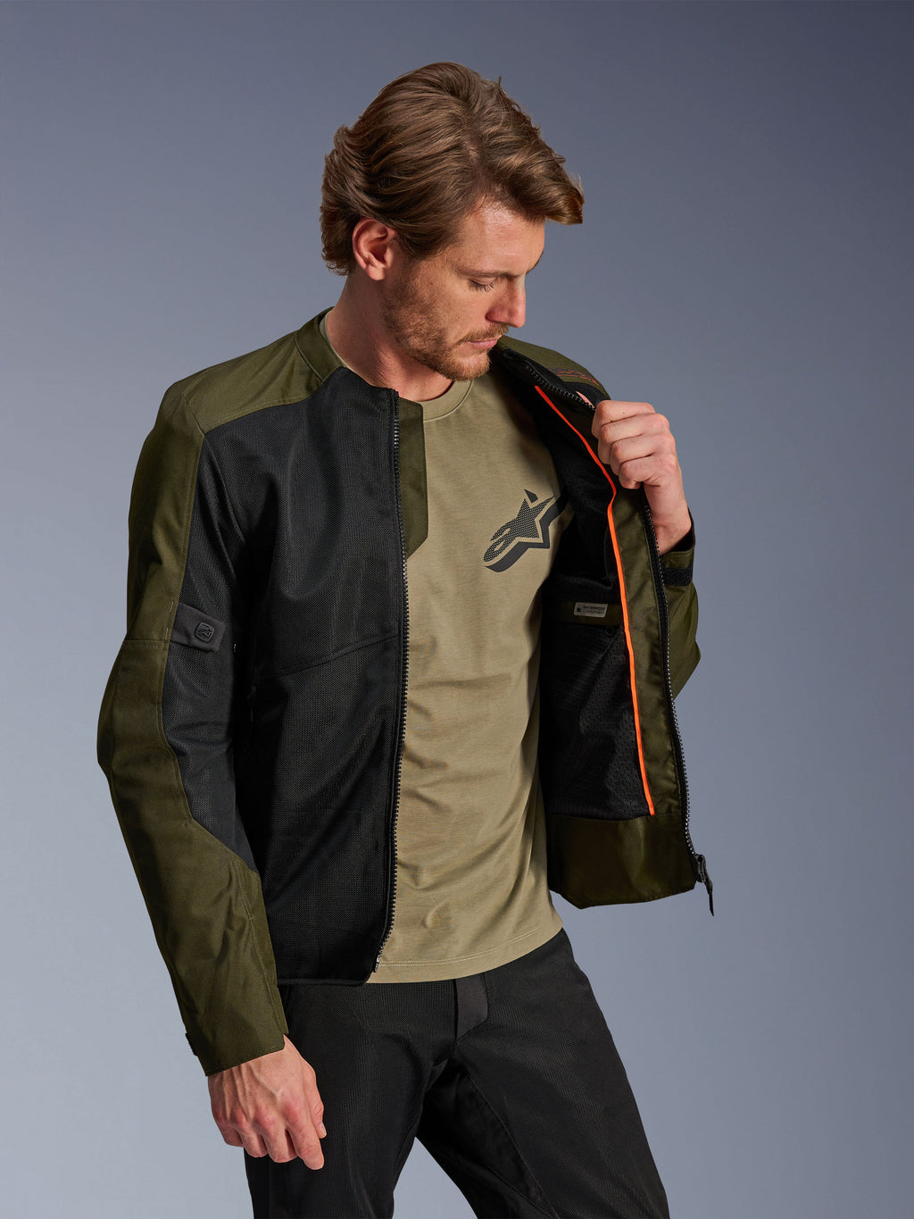 Alpinestars C-1 Air Jacke in Khaki und Schwarz, getragen von einem männlichen Model zur Darstellung des orangefarbenen Innenfutters und des wasserdichten Fachs, mit großen Tech-Mesh-Einsätzen für Luftzirkulation, urbaner zeitgenössischer Motorradstil kombiniert mit einem passenden olivfarbenen T-Shirt und einer schwarzen Hose