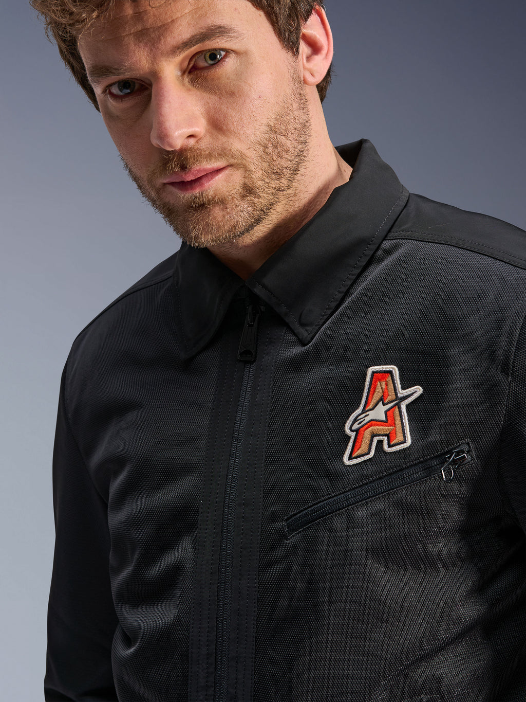 Alpinestars Ignite Air Jacke, Schwarz, mit traditionellem Hemdkragen und atmungsaktiver Mesh-Konstruktion, hervorgehoben durch einen rot-goldenen 'A'-Logo-Aufnäher auf der Brust, getragen von einem männlichen Model, urbane Motorradbekleidung