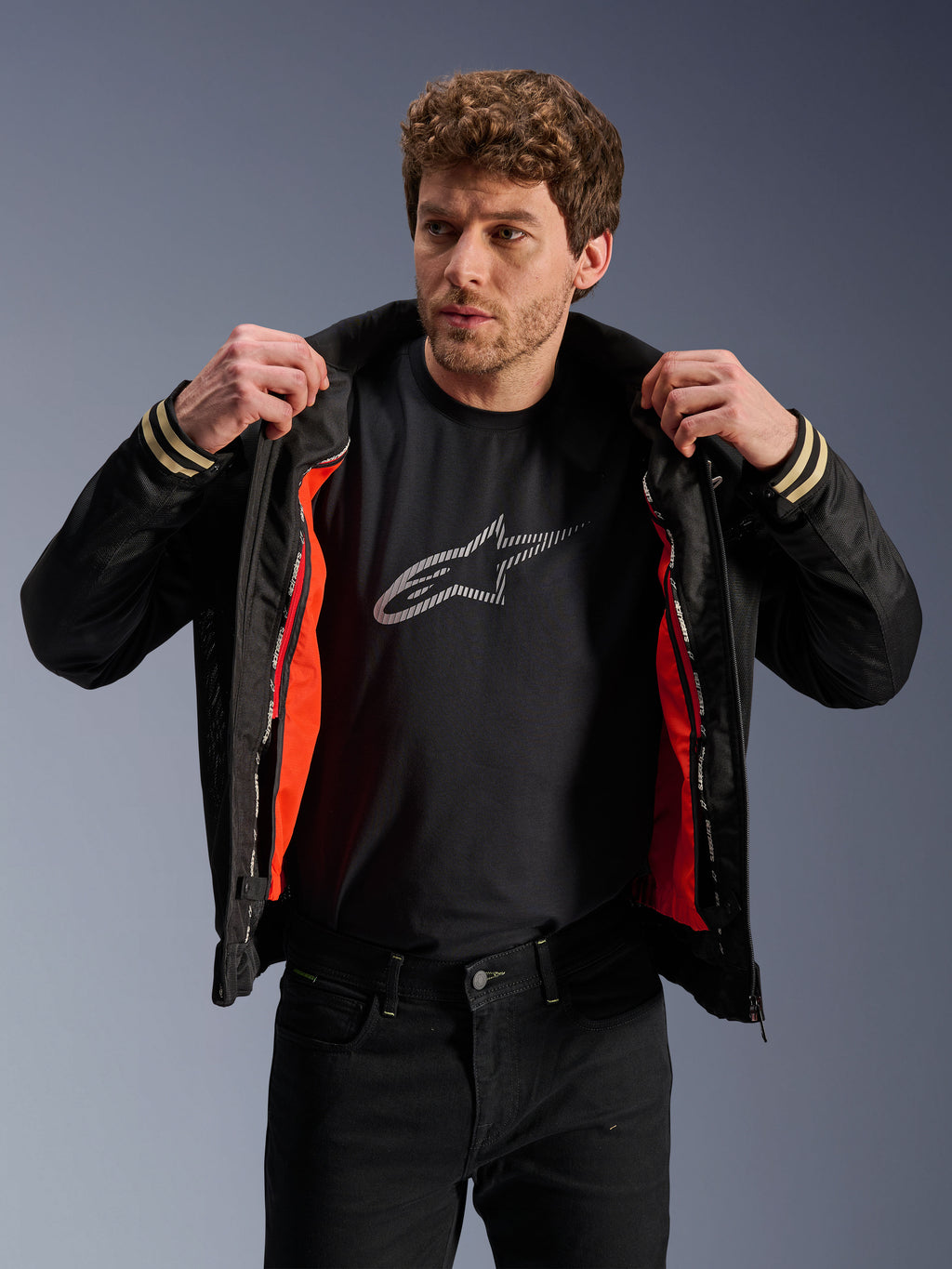 Alpinestars Ignite Air Jacke, Schwarz, mit Mesh-Körper und leuchtend orangefarbenem, abnehmbarem winddichtem Innenfutter, kombiniert mit einem schwarzen T-Shirt mit Astars-Logo und schwarzer Hose, urbaner zeitgenössischer Motorrad-Stil