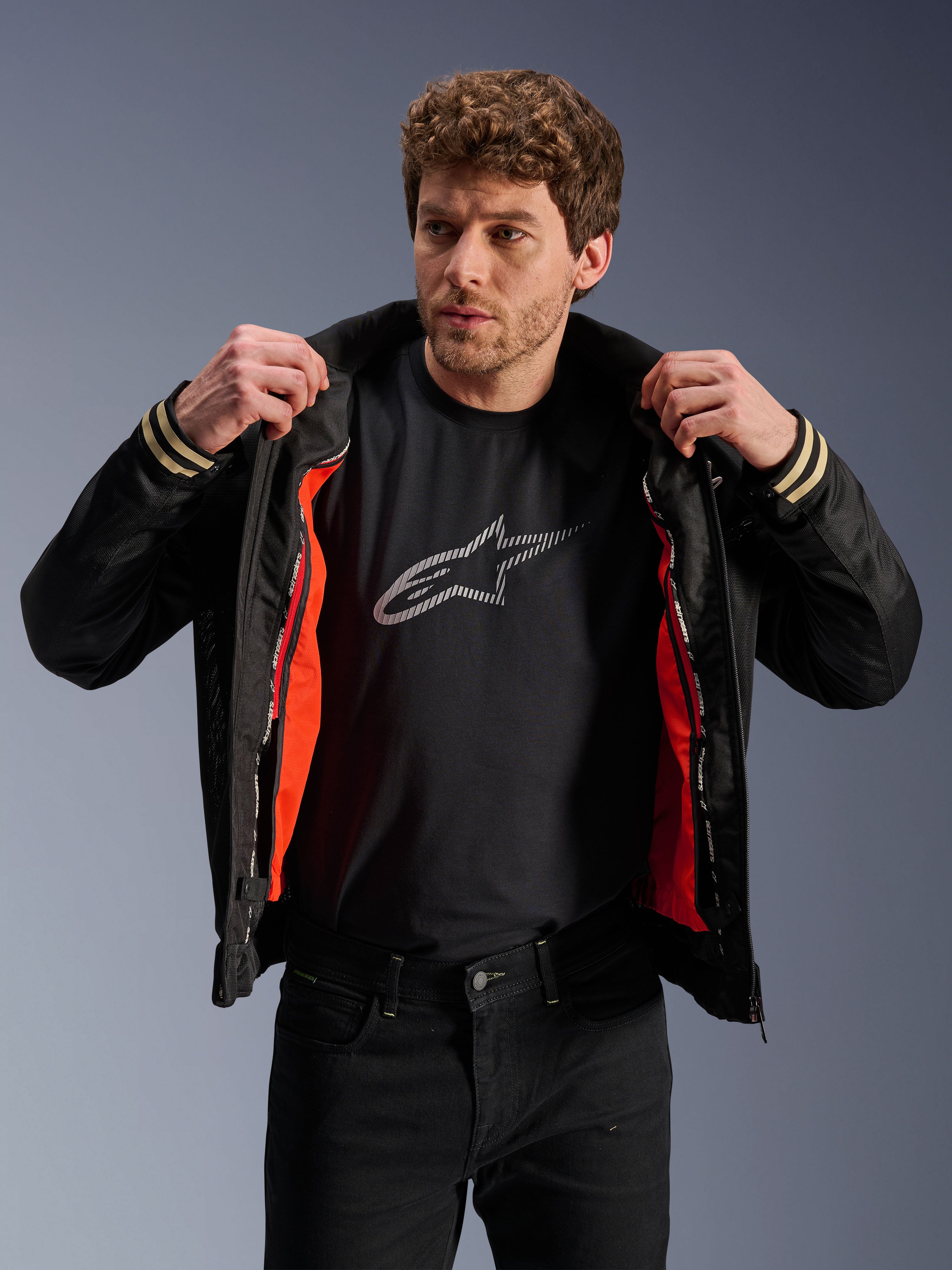 Alpinestars Ignite Air Jacke, Schwarz, mit Mesh-Körper und leuchtend orangefarbenem, abnehmbarem winddichtem Innenfutter, kombiniert mit einem schwarzen T-Shirt mit Astars-Logo und schwarzer Hose, urbaner zeitgenössischer Motorrad-Stil