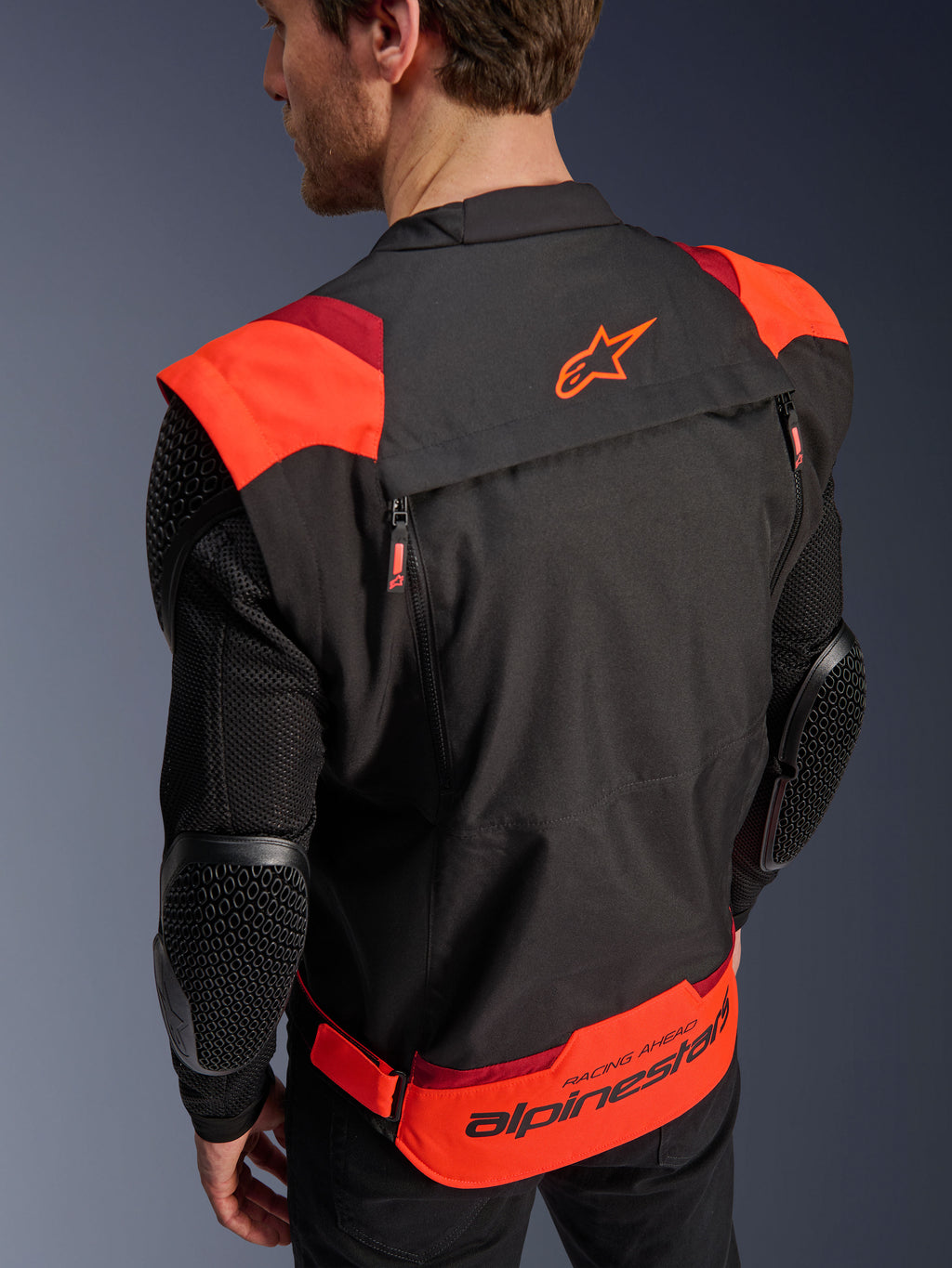 Alpinestars T-Stunt Air Jacke, Schwarz, Fluo-Rot und Dunkelrot, Rückansicht an einem Modell, zeigt Mesh-Ärmel mit wabenförmig strukturierten Ellbogenprotektoren, Alpinestars Stern-Logo auf dem oberen Rücken und markantes Branding am unteren Rücken mit 'RACING AHEAD'-Schriftzug, modulares Design mit sportlichem Schnitt für das Motorradfahren.