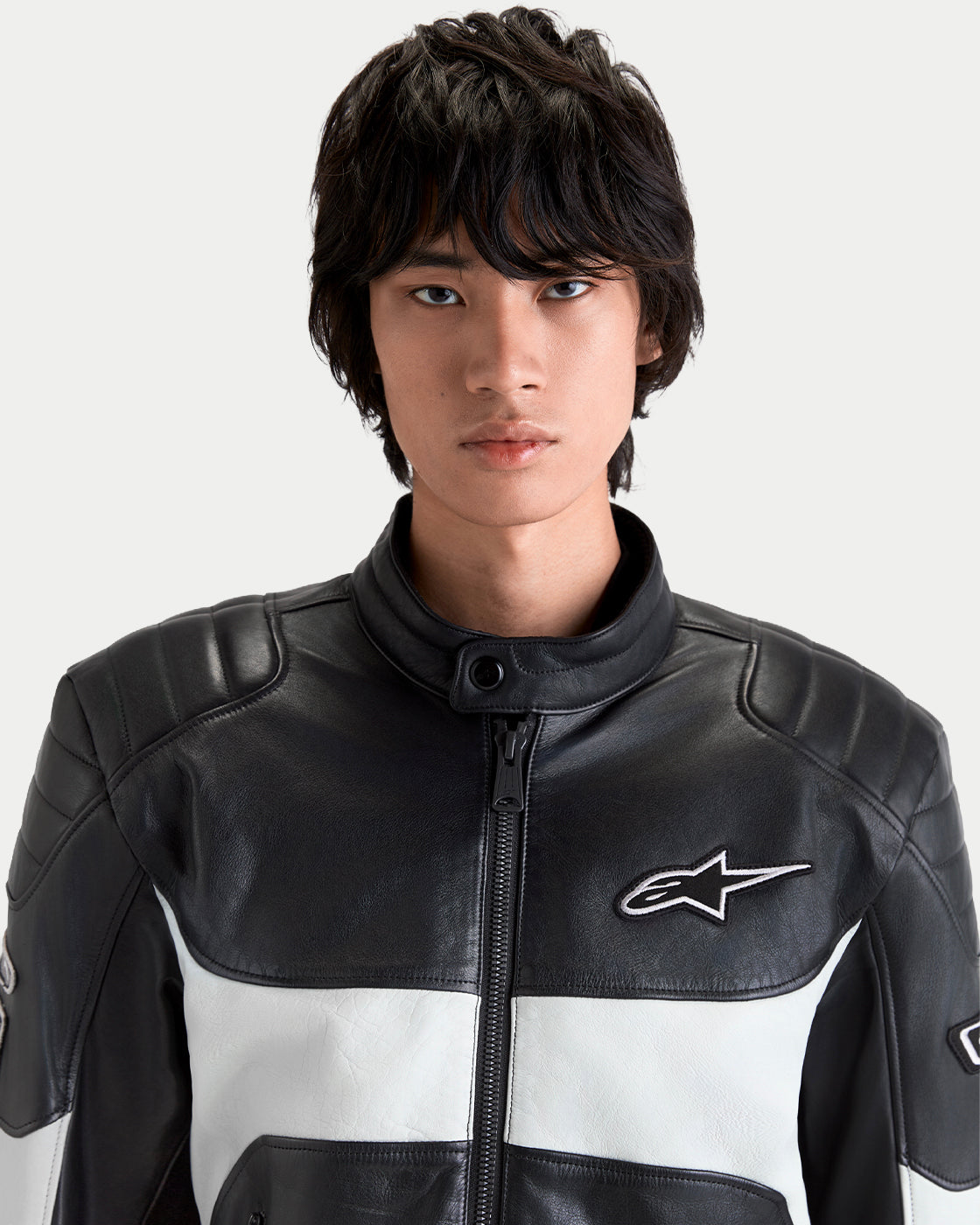 Alpinestars Revern Leather Jacket, men's motorcycle leather jacket in black and white, close-up portrait of a model wearing the riding jacket featuring shoulder padding, high collar, and the Alpinestars logo on the chest