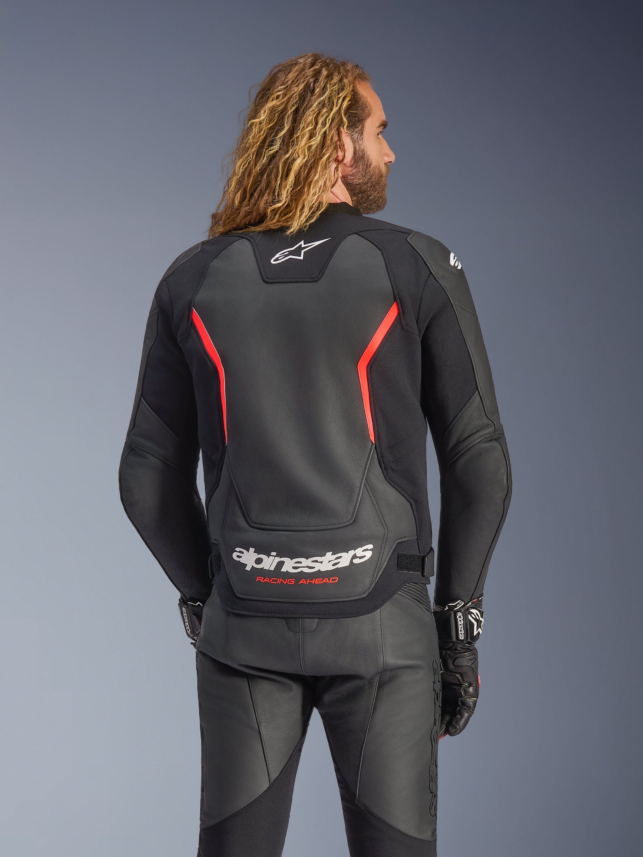 Alpinestars GP Force V2 Lederjacke, Schwarz und Fluo-Rot, Rückansicht an einem Model, mit weißem Alpinestars-Logo und „RACING AHEAD“-Schriftzug, kombiniert mit schwarzer Leder-Motorradhose und Handschuhen.
