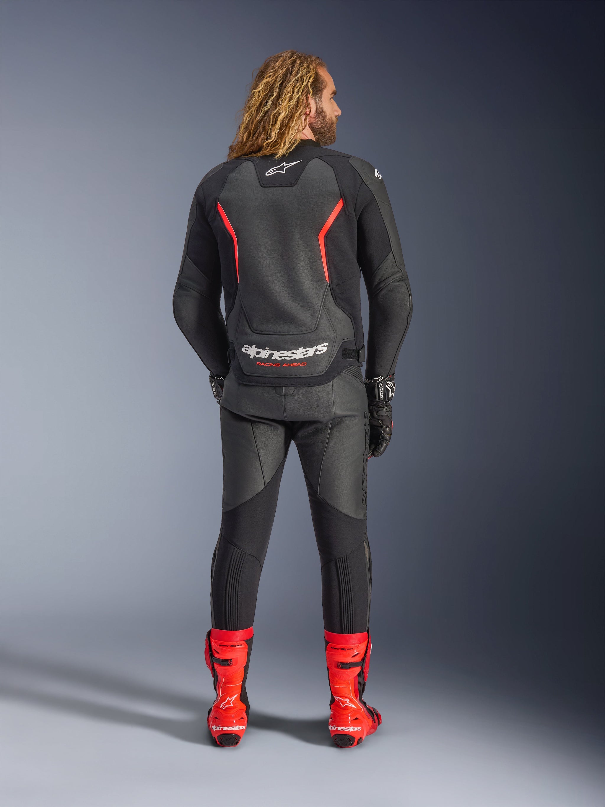 Alpinestars GP Force V2 Lederjacke, Schwarz und Fluo-Rot, Rückansicht, getragen mit passender schwarzer Lederhose und leuchtend roten Rennstiefeln, mit roten Akzentlinien und markantem weißen Alpinestars-Branding auf dem Rücken, professionelles Sport-Motorrad-Outfit.