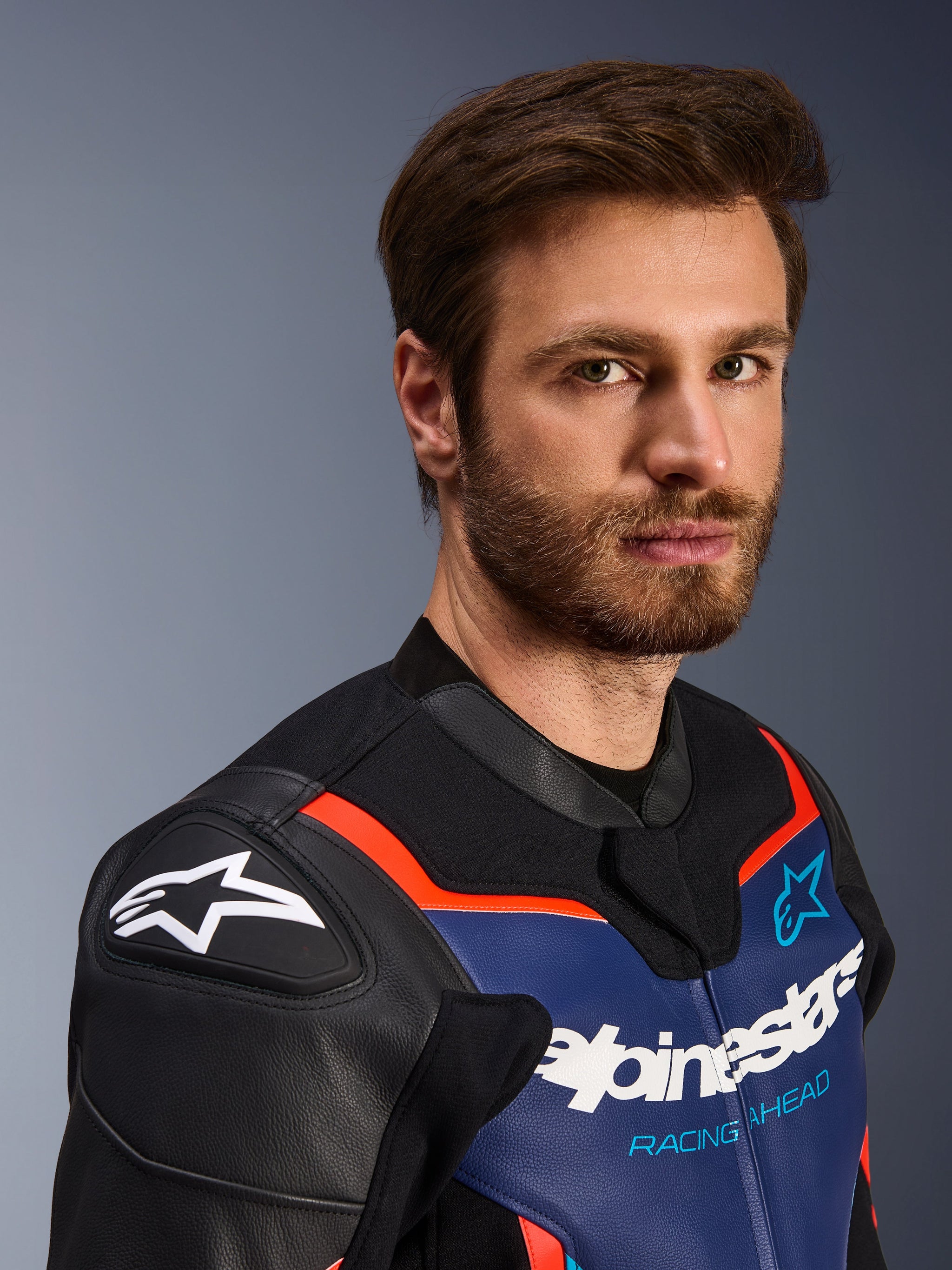 Alpinestars GP Force V2 Lederjacke, Schwarz, Blau und Fluo-Rot, Nahaufnahme eines männlichen Fahrers, mit weißem Alpinestars-Logo auf schwarzen DFS-Schulterschutzschilden, blauem Leder-Brustpanel mit weißem Branding und „Racing Ahead“-Schriftzug, fluo-roten Akzenten, Premium-Sport-Motorradausrüstung.
