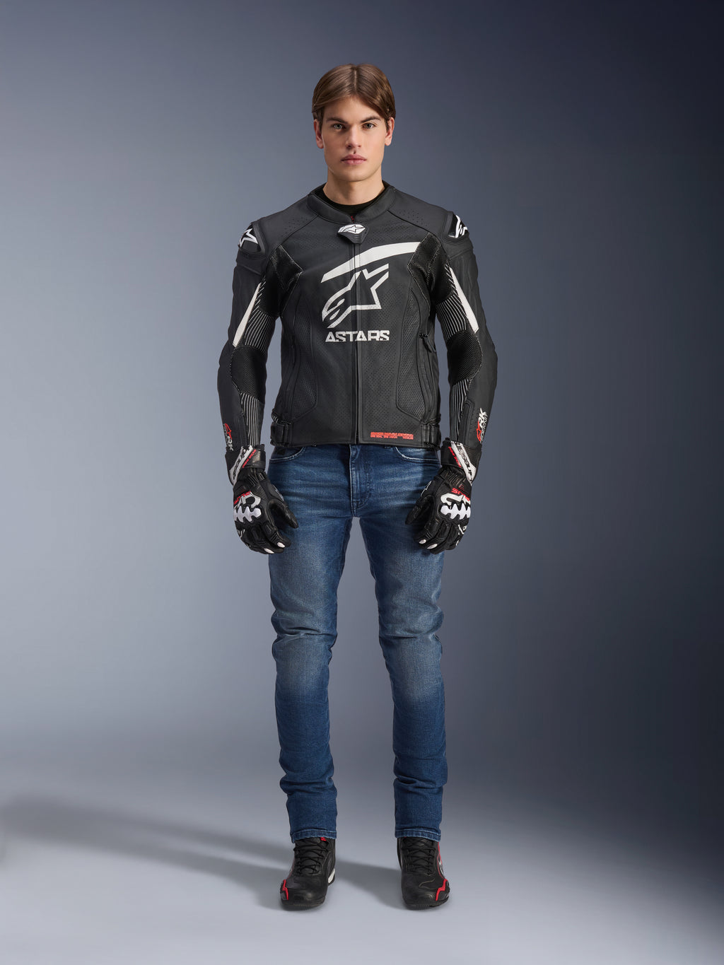 Männliches Model in Alpinestars GP Plus R V4 Rideknit Lederjacke in Schwarz und Weiß, kombiniert mit blauen Motorradjeans, schwarzen Motorradhandschuhen und schwarzen Motorradschuhen, großes weißes Astars-Logo auf der Brust, perforierte Lederbahnen und weiße Rennstreifen an den Ärmeln, Ganzkörperansicht im Stehen