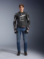 Männliches Model in Alpinestars GP Plus R V4 Rideknit Lederjacke in Schwarz und Weiß, kombiniert mit blauen Motorradjeans, schwarzen Motorradhandschuhen und schwarzen Motorradschuhen, großes weißes Astars-Logo auf der Brust, perforierte Lederbahnen und weiße Rennstreifen an den Ärmeln, Ganzkörperansicht im Stehen