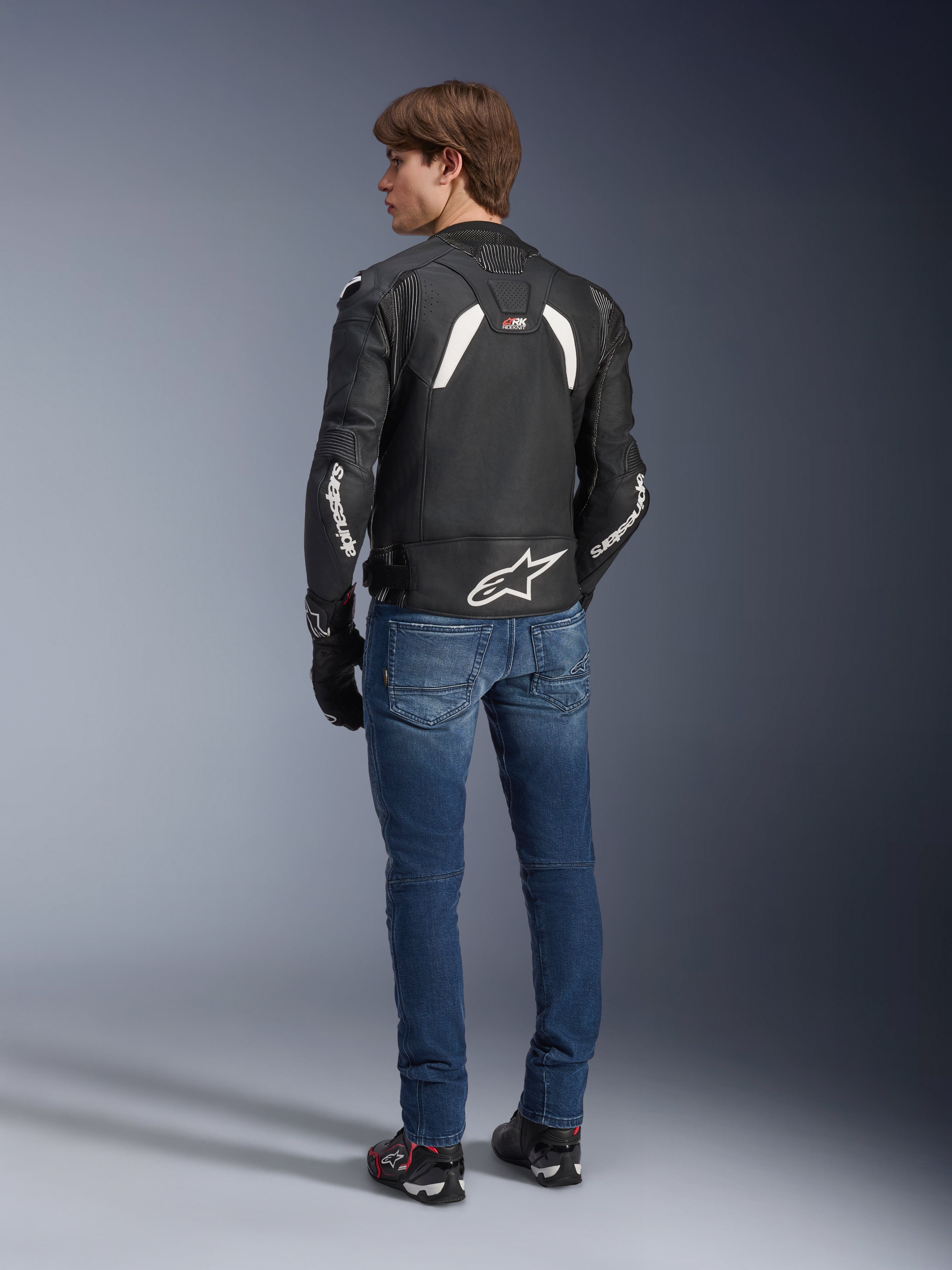 Alpinestars GP Plus R V4 Rideknit Lederjacke in Schwarz-Weiß, Rückansicht an einem Model, mit weißem Alpinestars-Branding an den Ärmeln und einem großen Stern-Logo auf dem unteren Rücken, kombiniert mit blauen Jeans und schwarzen Motorradschuhen, technische Motorradausrüstung.