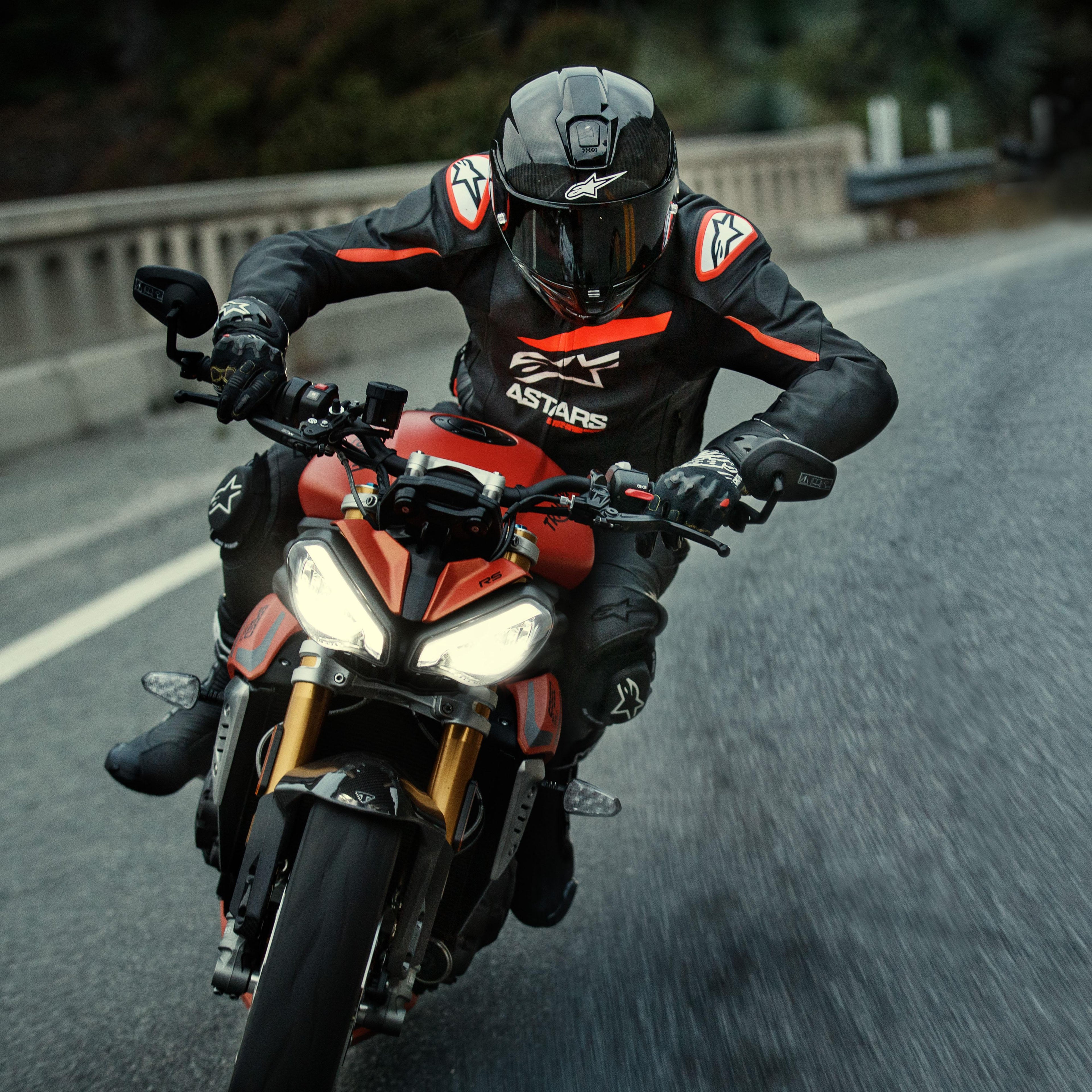 Motorradfahrer auf einem roten Sportmotorrad, Alpinestars GP Plus R V4 Rideknit Lederjacke, Schwarz mit fluoroten und weißen Details, mit weißem Alpinestars-Logo und „ASTARS“-Schriftzug, kombiniert mit schwarzem Rennhelm und Handschuhen, Performance-Fahrausrüstung in Aktion auf einer Straße.