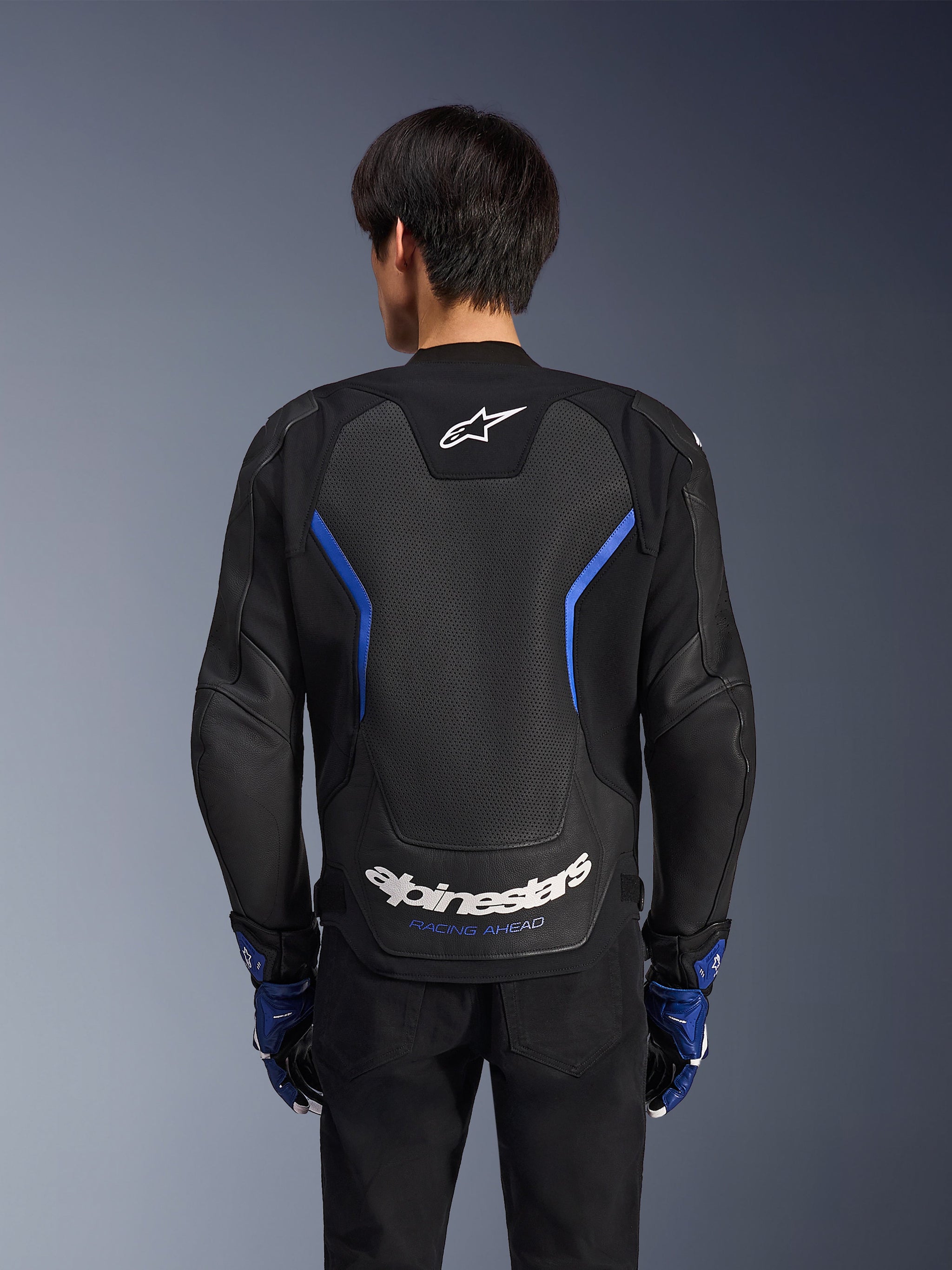 Alpinestars GP Force V2 Airflow Lederjacke, Schwarz Metallic-Blau, Rückansicht, mit einem großen perforierten Rückenpanel zur Belüftung, blauen Akzenten und weißem Alpinestars-Branding, getragen mit passenden blauen Handschuhen und schwarzer Hose für sportliches Fahren.