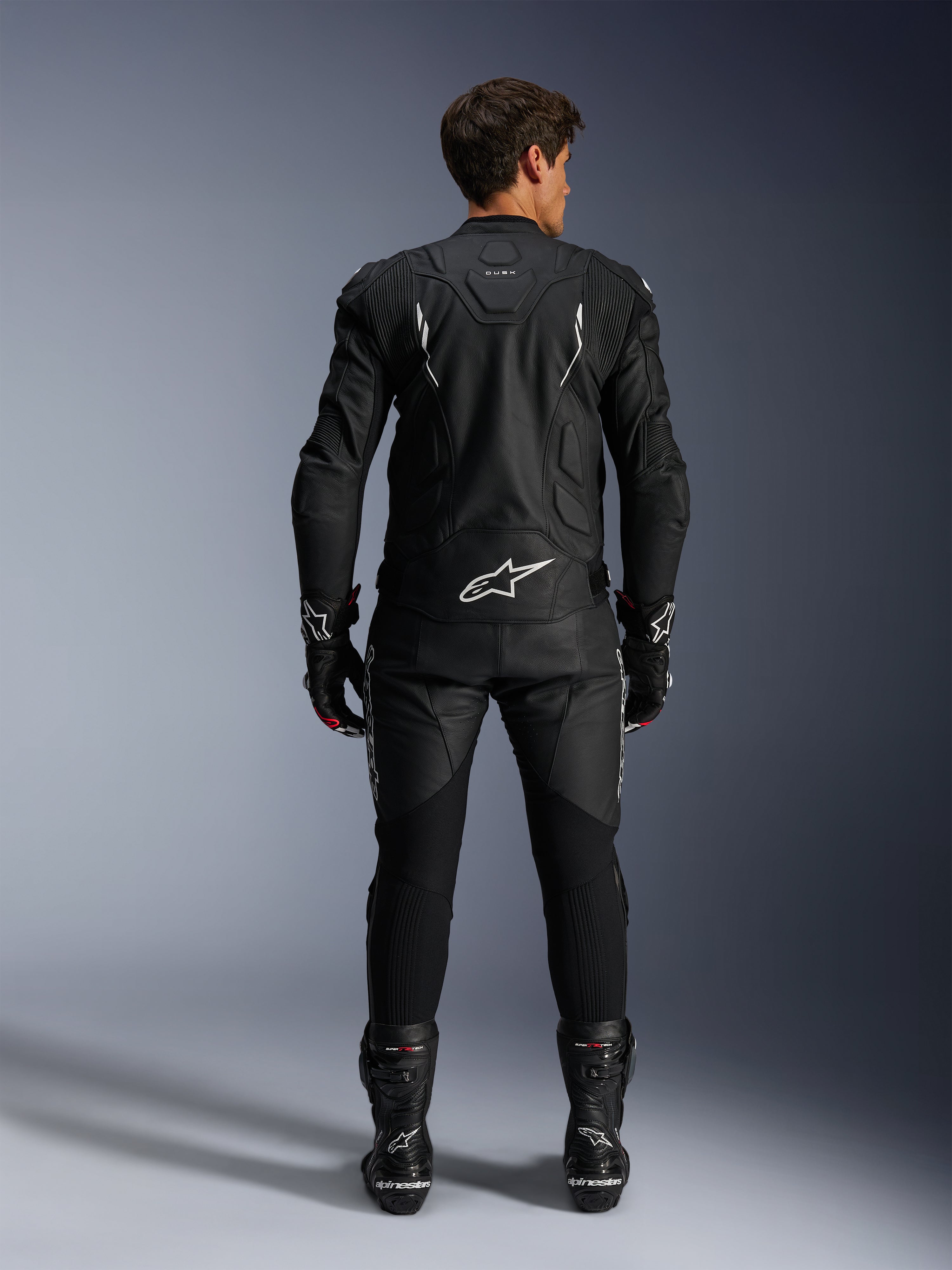 Motorradfahrer in der Rückansicht mit der Alpinestars Dusk Lederjacke und der Missile V3 Lederhose, kompletter schwarzer Lederanzug mit weißen Akzenten, Jacke mit „Dusk“-Branding und Alpinestars-Logo auf dem Rücken, kombiniert mit schwarzen Rennstiefeln und Handschuhen, abgestimmtes Motorsport-Outfit für den Straßenrennsport.