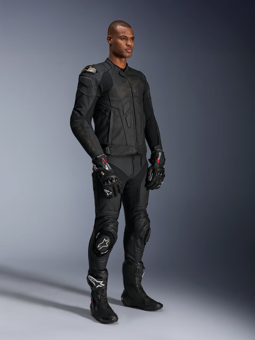 Male model wearing the Alpinestars Dusk Leather Jacket in Black Laser, paired with matching black leather racing pants, GP Pro gloves, and performance track boots. Full motorcycle racing gear with external DFS shoulder protection and a sleek athletic fit for sport and road riding.