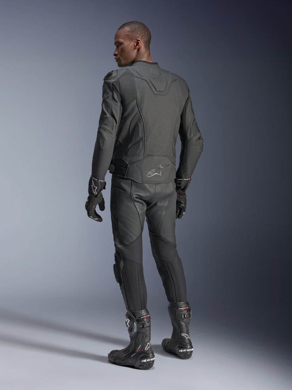 Männlicher Fahrer von hinten in der Alpinestars Dusk Airflow Lederjacke in Black Black, kombiniert mit passender Lederhose, Handschuhen und Stiefeln, hebt die umfassenden Perforationen am Rücken und die sportliche Performance-Passform hervor.