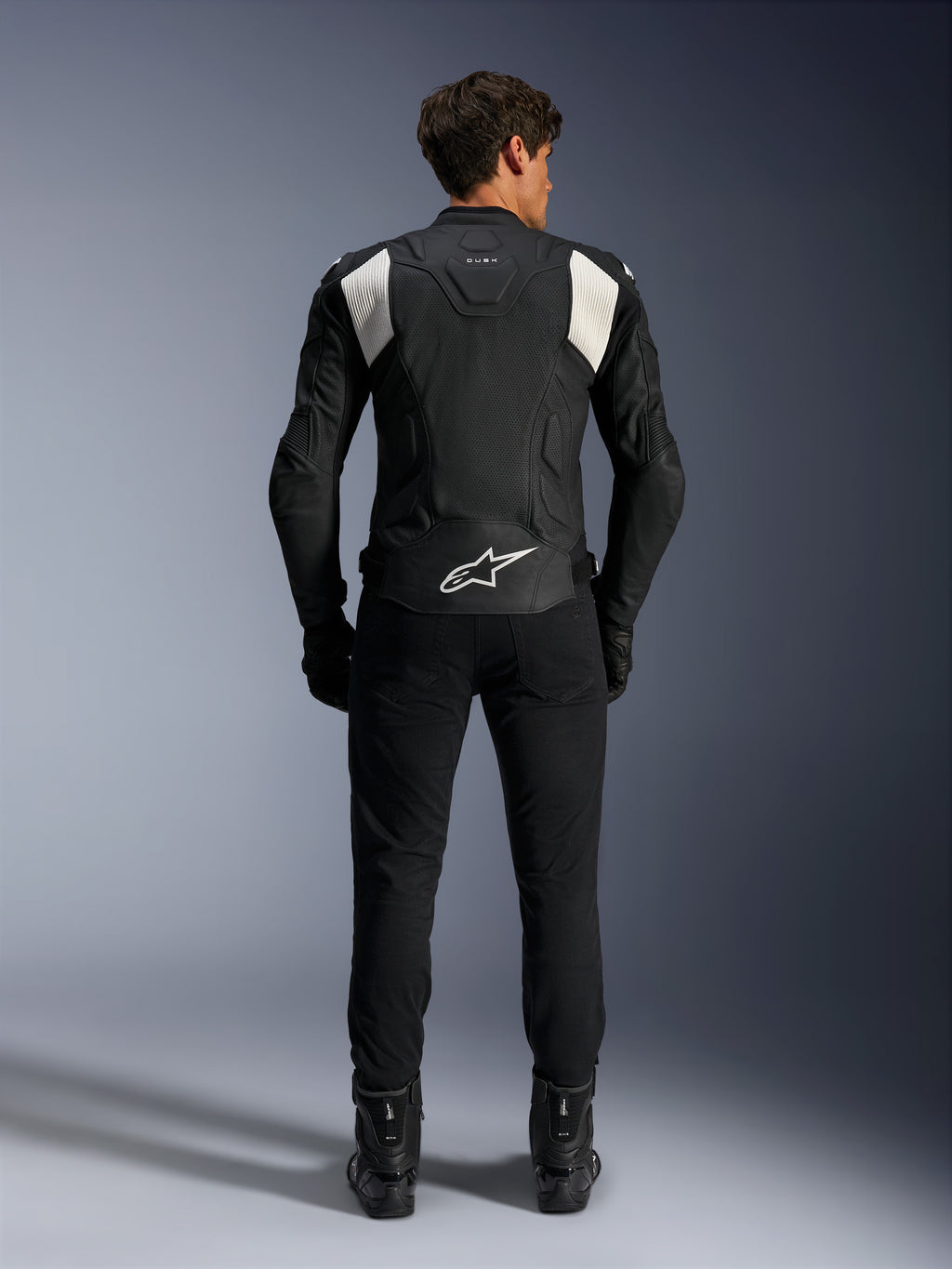 Alpinestars Dusk Airflow Lederjacke, Schwarz und Weiß, Rückansicht eines Fahrers mit schwarzer Hose und Stiefeln, mit perforiertem Leder für Belüftung, weißen Akkordeon-Stretcheinsätzen an den Schultern und weißem Branding am unteren Rücken