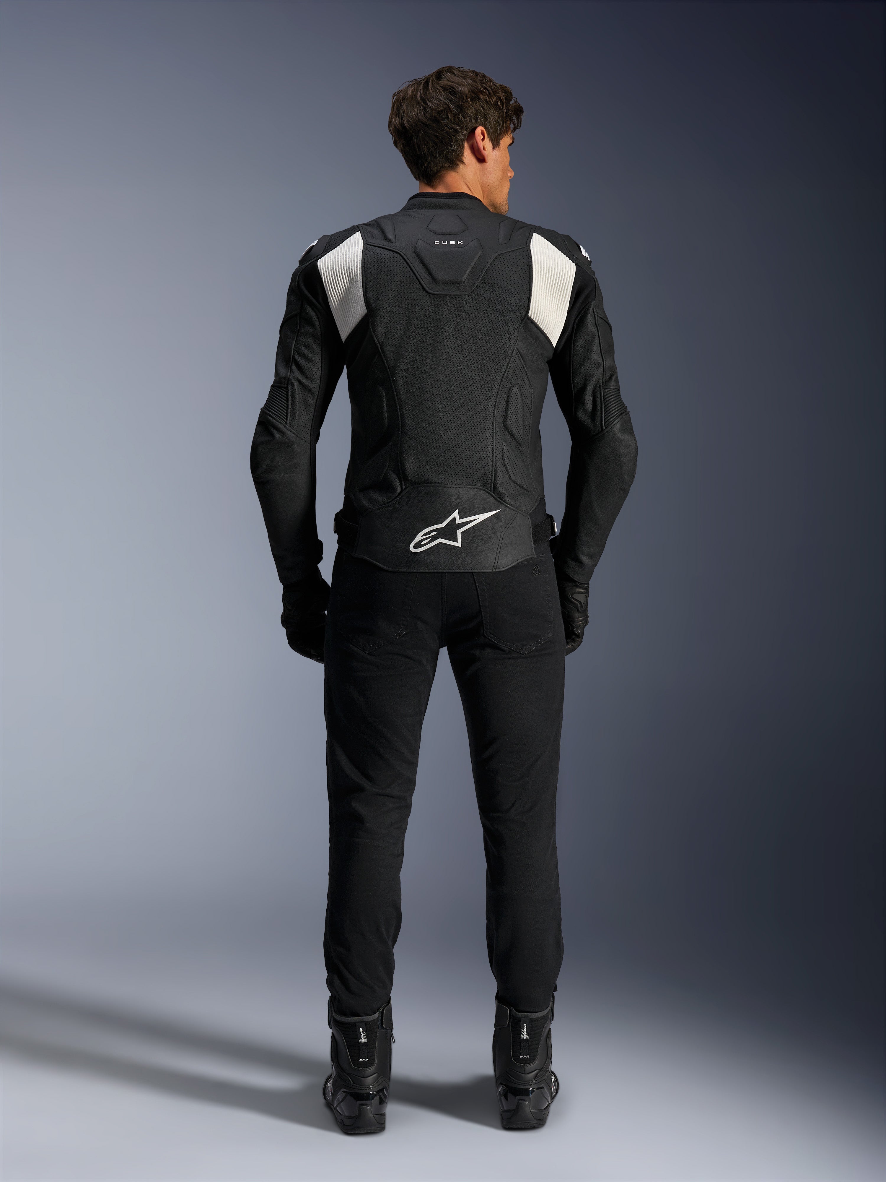 Alpinestars Dusk Airflow Lederjacke, Schwarz und Weiß, Rückansicht eines Fahrers mit schwarzer Hose und Stiefeln, mit perforiertem Leder für Belüftung, weißen Akkordeon-Stretcheinsätzen an den Schultern und weißem Branding am unteren Rücken