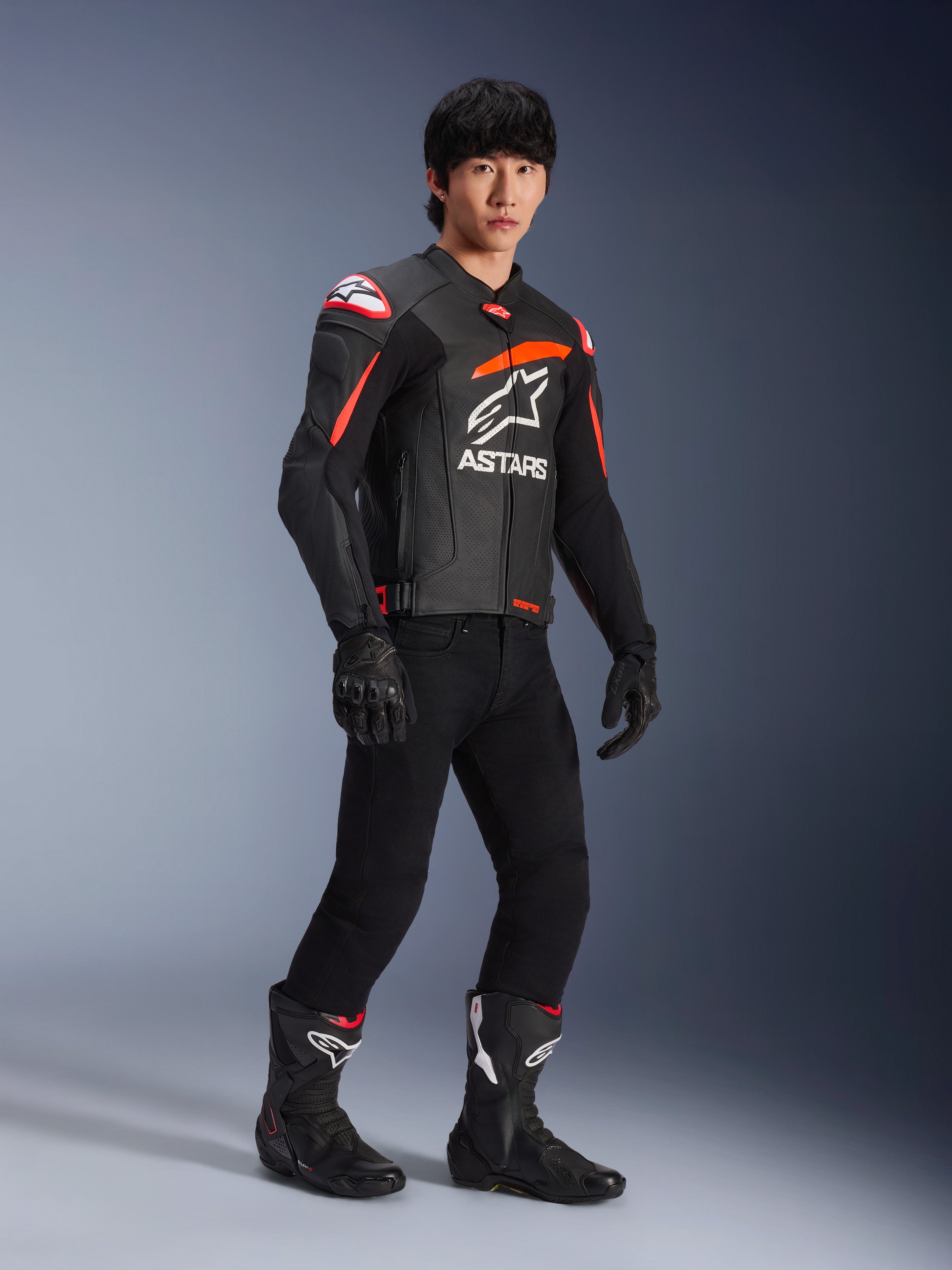Male rider wearing Alpinestars GP Plus R V4 Airflow Leather Jacket, Black with Red Fluo and White accents, featuring perforated leather and ASTARS chest logo, worn with black riding pants and black motorcycle boots, sport performance gear