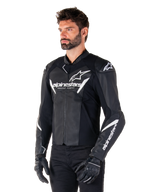 Alpinestars Faster V3 Lederjacke, Sport-Motorradjacke, Schwarz und Weiß, auffälliges weißes Alpinestars-Logo und 'Racing Ahead'-Schriftzug auf der Brust, Stern-Logos auf den Schultern, hochwertige Lederkonstruktion mit Stretcheinsätzen, getragen von einem Modell mit schwarzer Hose und Handschuhen