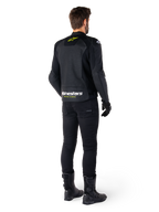 Rückansicht eines Fahrers in der Alpinestars Faster V3 Airflow Lederjacke, Schwarz mit weißen und neongelben Akzenten, kombiniert mit schwarzer Hose und Stiefeln, mit perforiertem Leder zur Belüftung und markantem „RACING AHEAD“-Branding am unteren Rücken.