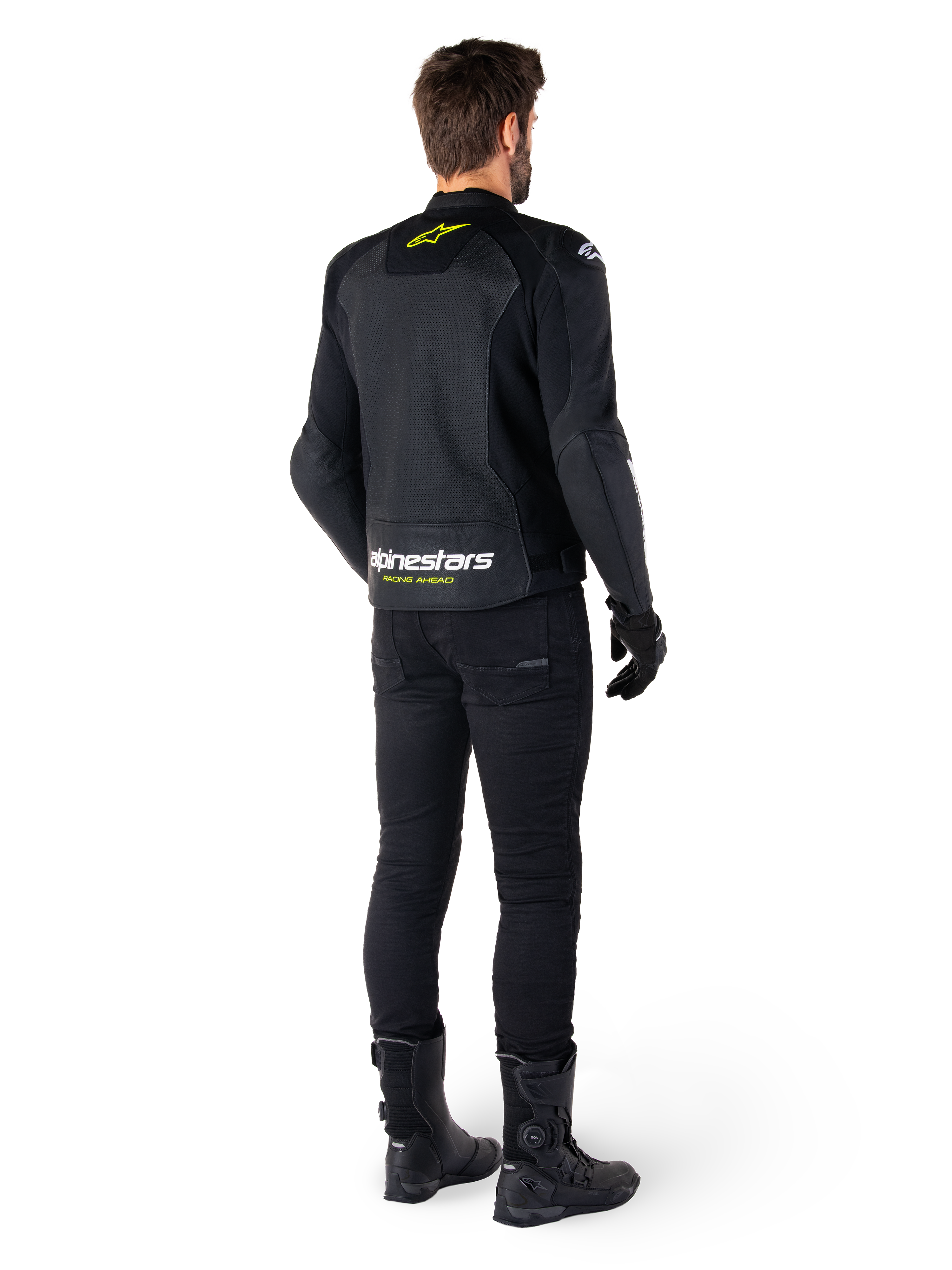 Rückansicht eines Fahrers in der Alpinestars Faster V3 Airflow Lederjacke, Schwarz mit weißen und neongelben Akzenten, kombiniert mit schwarzer Hose und Stiefeln, mit perforiertem Leder zur Belüftung und markantem „RACING AHEAD“-Branding am unteren Rücken.