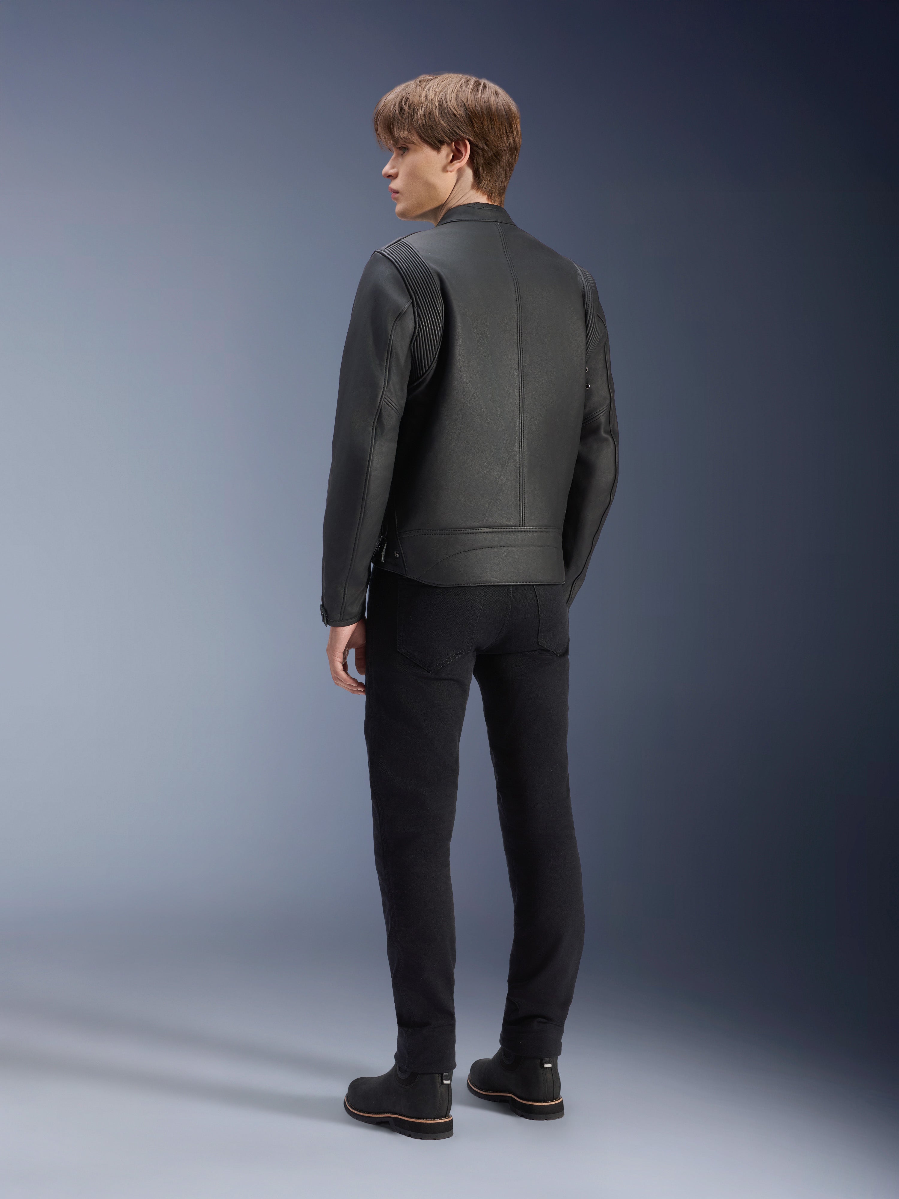 Alpinestars Dyno Lederjacke, Schwarz, Rückansicht an einem männlichen Model, mit gerippten Schultereinsätzen, kombiniert mit schwarzer Hose und Stiefeln, Heritage-inspirierter urbaner Motorradstil
