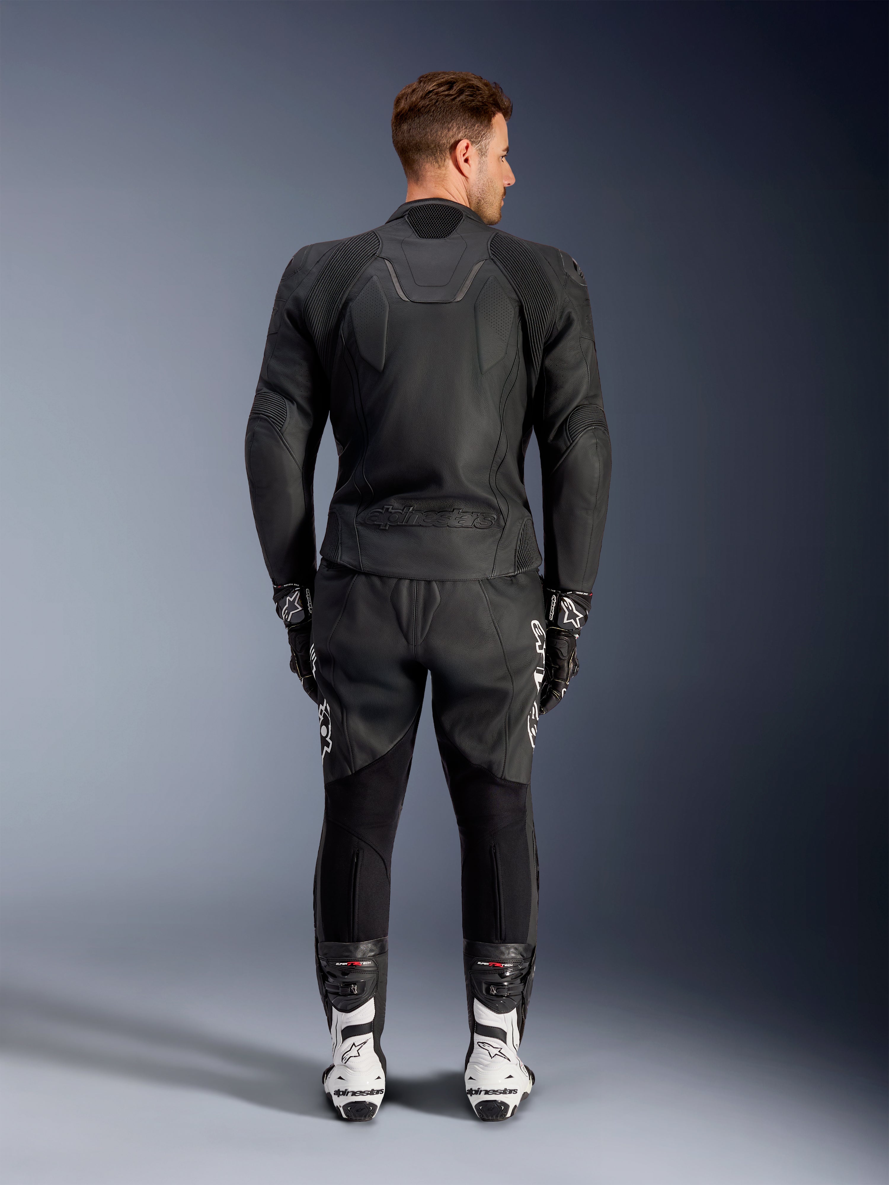 Rückansicht eines Mannes in der Alpinestars Caliber Lederjacke in Schwarz, mit geprägtem Logo und perforierten Lederbahnen, kombiniert mit einer schwarzen Motorrad-Lederhose, schwarzen Handschuhen und weißen Rennstiefeln.