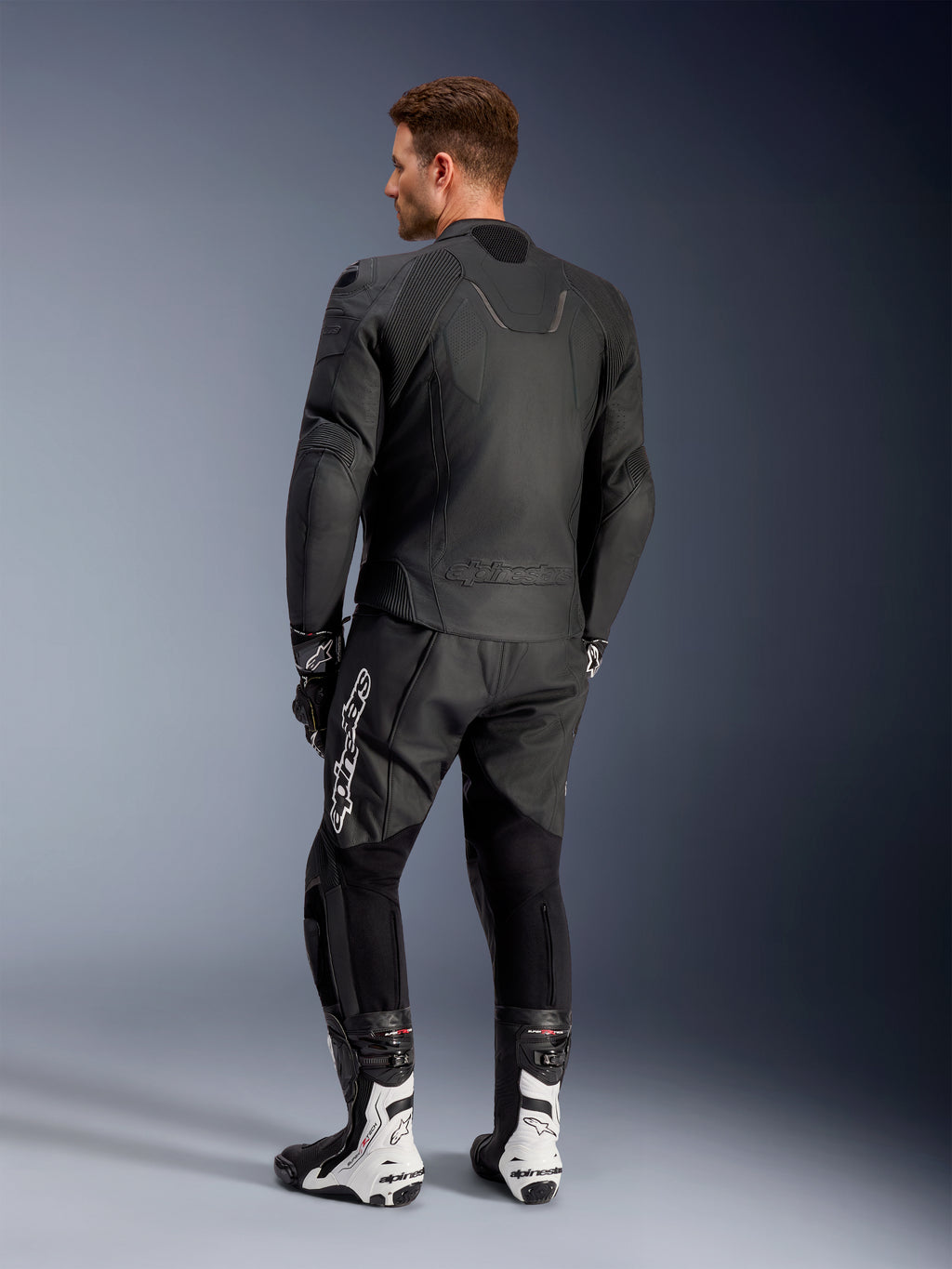 Alpinestars Caliber Lederjacke, schwarze Motorradjacke, Rückansicht, mit perforiertem Leder und Akkordeon-Stretcheinsätzen an Taille und Schultern, geprägtes Logo, getragen mit schwarzer Lederhose und weißen Rennstiefeln
