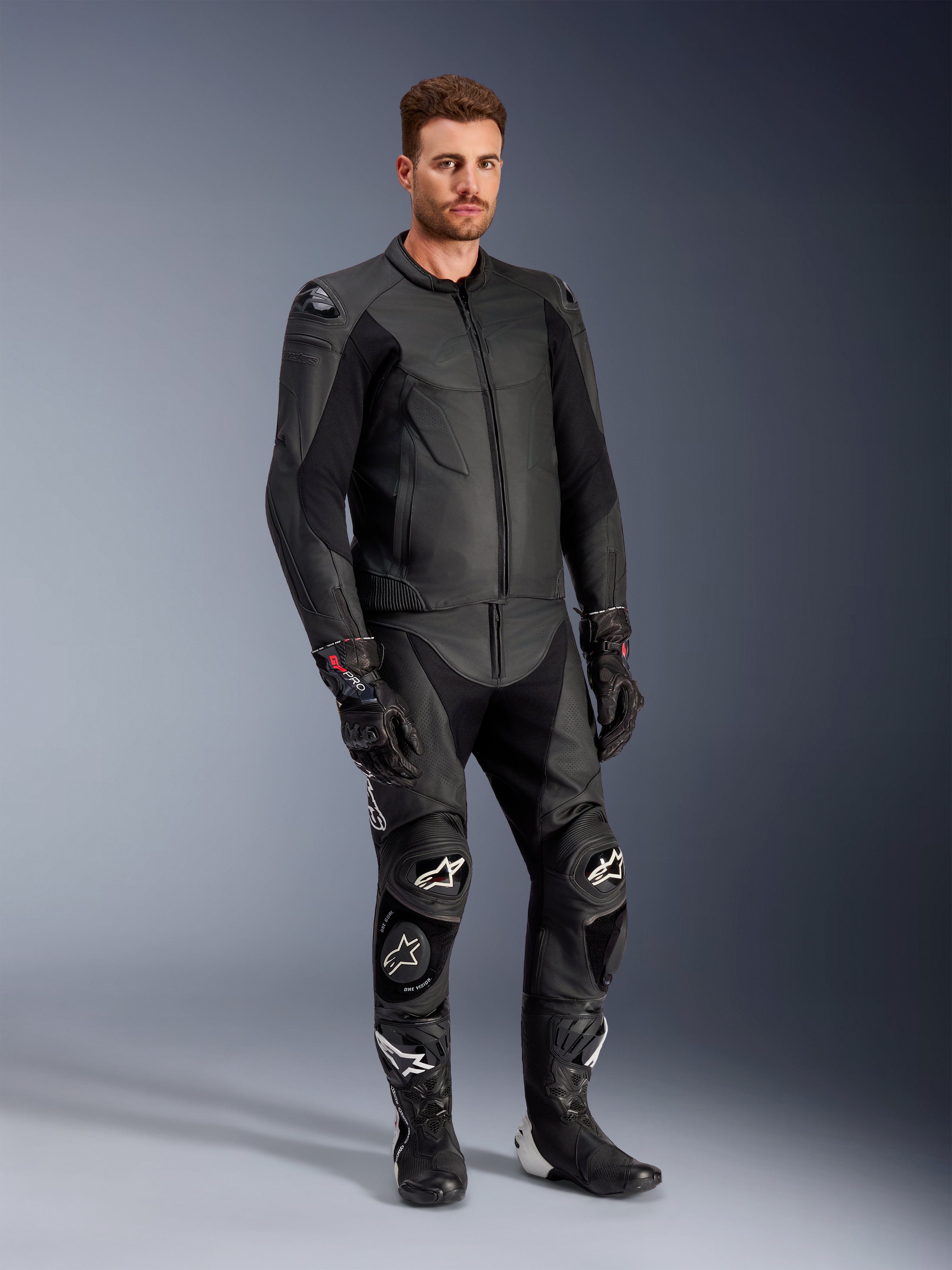 Männliches Modell in der Alpinestars Caliber Lederjacke in Schwarz, kombiniert mit passender schwarzer Leder-Motorradhose, Rennhandschuhen und Stiefeln, präsentiert ein komplettes Sport-Road-Outfit.