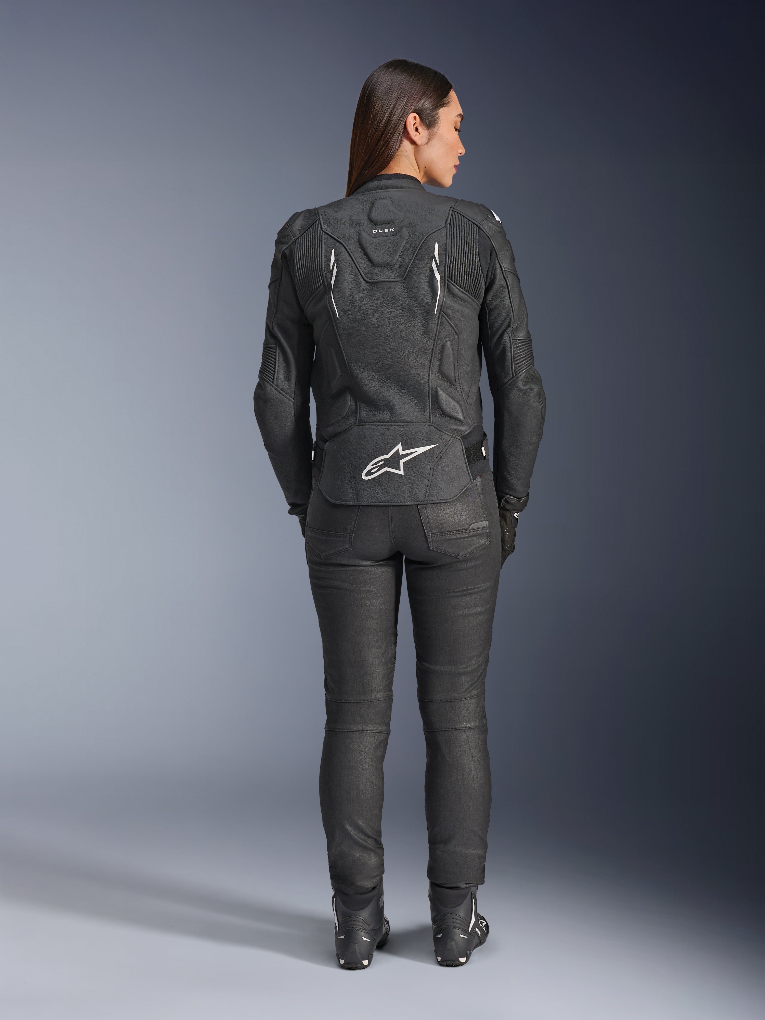 Alpinestars Woman Stella Dusk Leather Jacket, Black, Rear view featuring white Alpinestars logo on the lower back and 'DUSK' branding on the upper back, worn with black riding pants and boots, showcasing the contoured women's fit and protective leather construction.