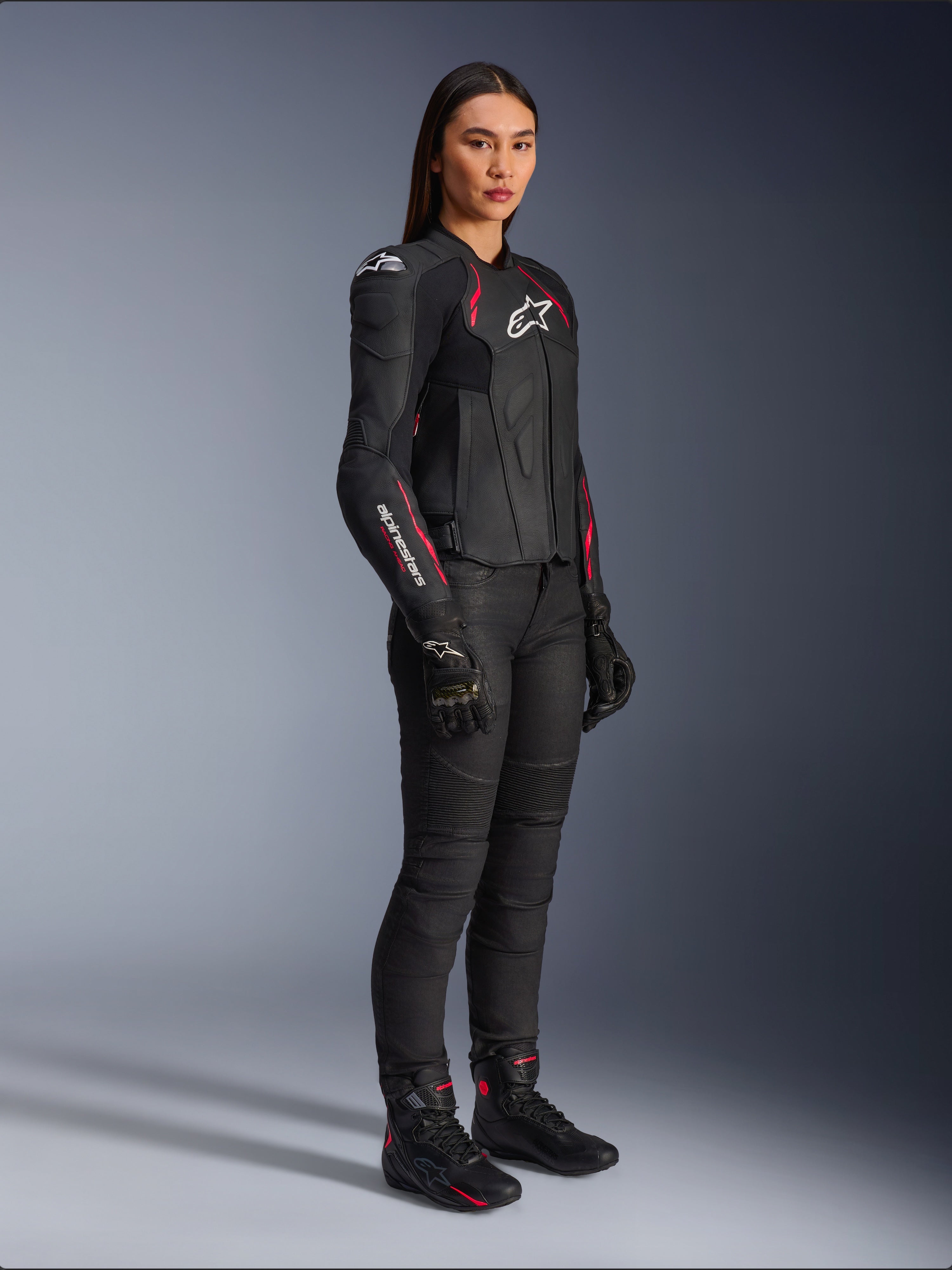 Alpinestars Stella Dusk Damen-Lederjacke, Schwarz, Diva-Pink und Weiß, getragen von einem weiblichen Model mit schwarzer Motorradhose und Stiefeln, elegante Lederkonstruktion mit pinkfarbenen Details und weißem Branding