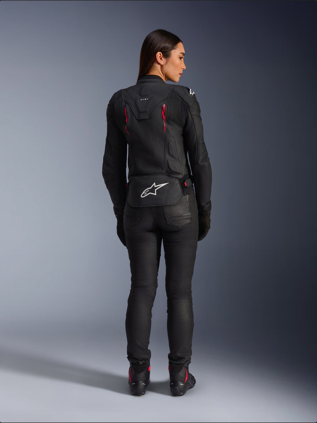 Alpinestars Stella Dusk Lederjacke, Rückansicht, schwarzes Leder mit Akzenten in Diva Pink und Weiß, weißes Alpinestars Logo am unteren Rücken und pinke Belüftungsdetails, getragen mit schwarzer Motorradhose und Stiefeln, hochwertige Sport-Motorradausrüstung für Damen